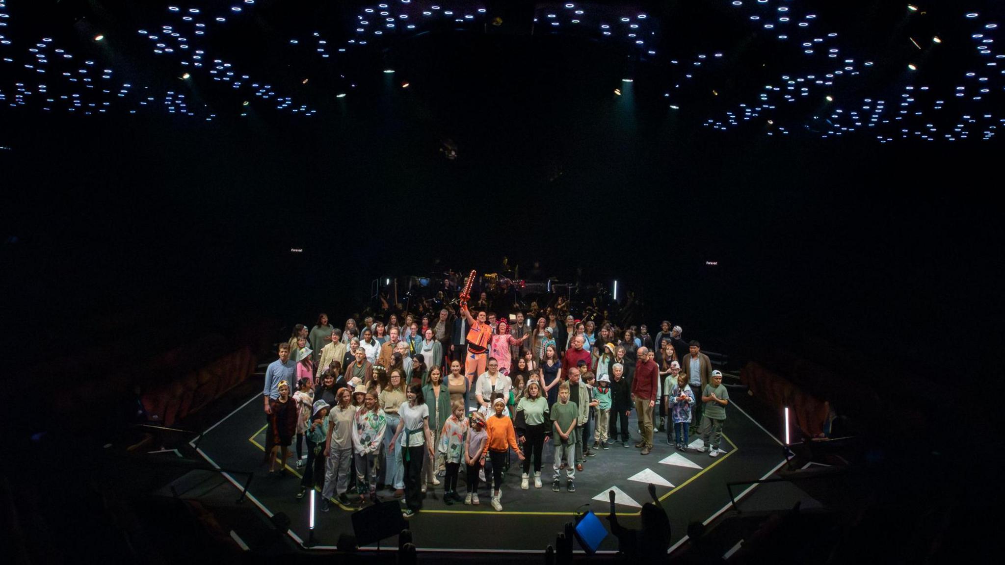 A view of the Crucible mainstage shows around 50 people looking out into the audience, with a man holding a sword aloft at their centre