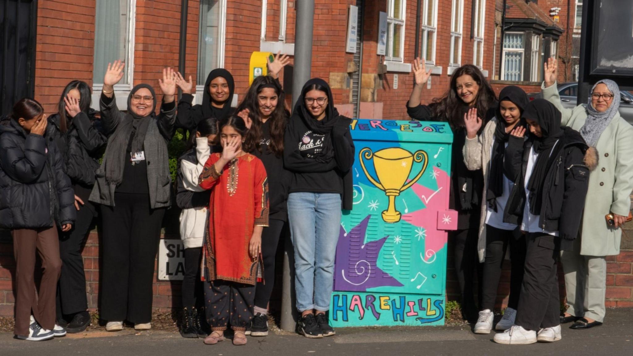 Eleven girls stand in a line near a street electrical box which has been painted with colourful letters reading "Heroes of Harehills". To the right of the girls is Nahid Rasool, who has worked at the centre since it began. The centre, a simple brick building, is behind the group of people.