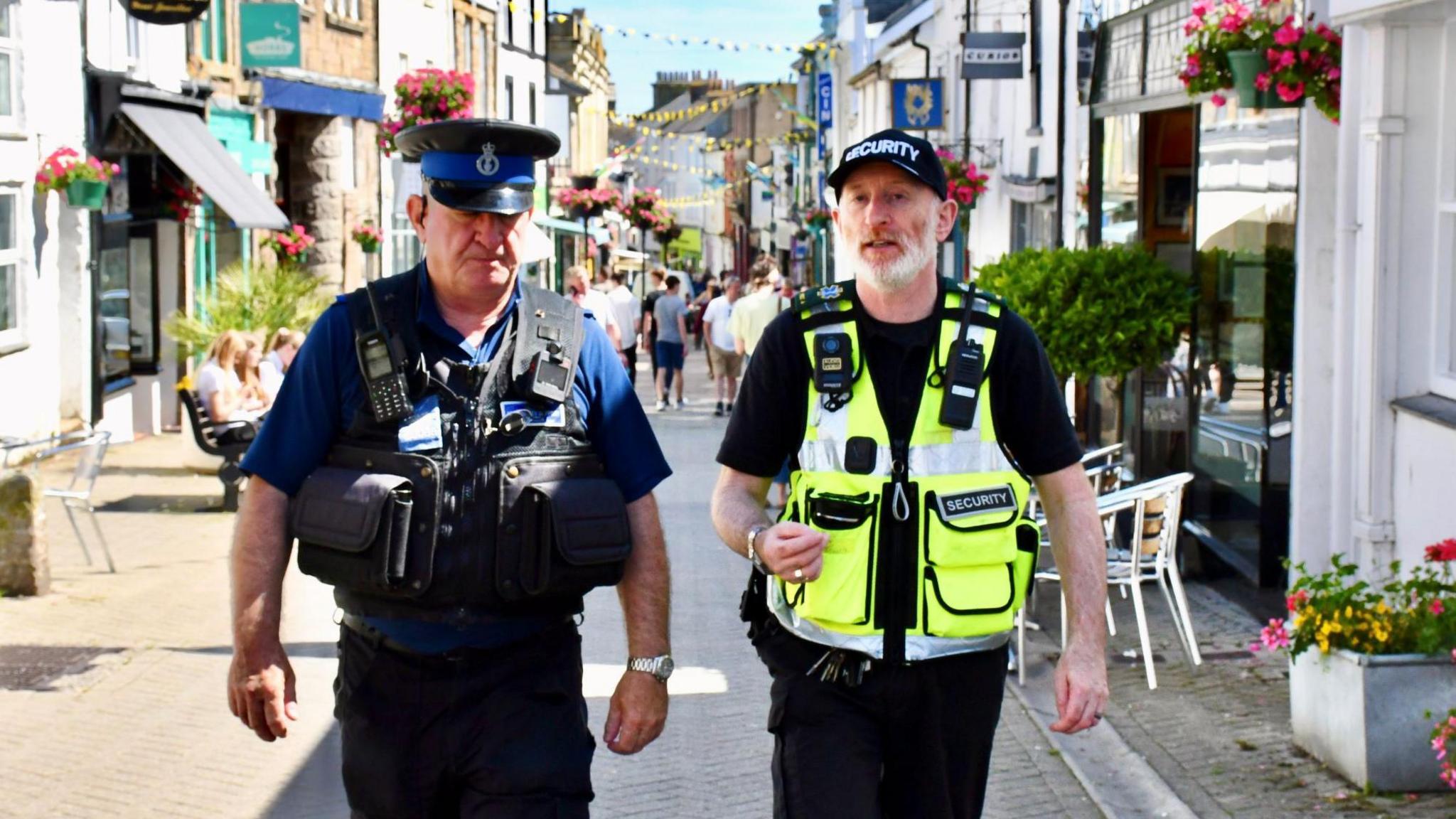 A police community support officer and a security guard walking down a road in Penzance on a sunny day.