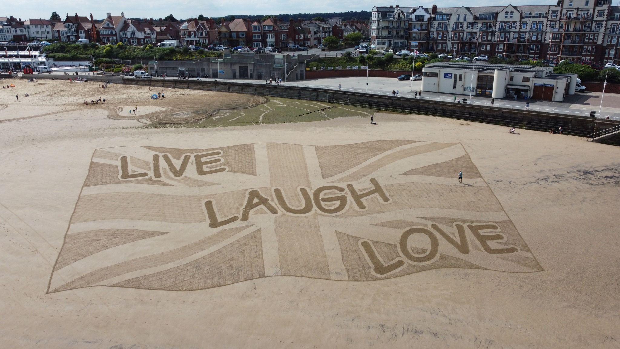 A large sand artwork on Bridlington beach, with a Union Jack flag and the slogan live, laugh, love, in a comic sans font.