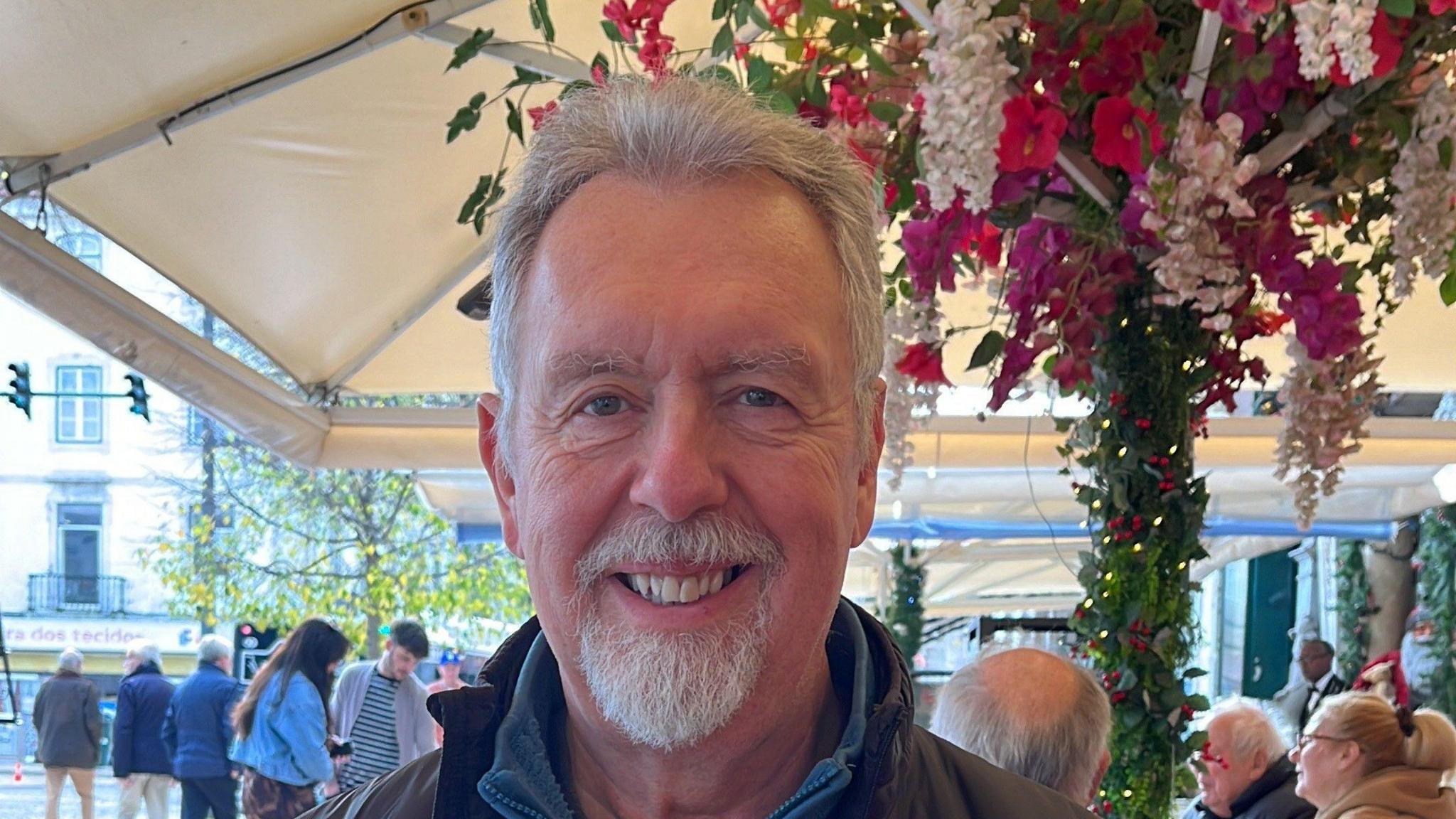 Peter Roberts, with grey hair and a grey goatee sat in a busy restaurant smiling at the camera