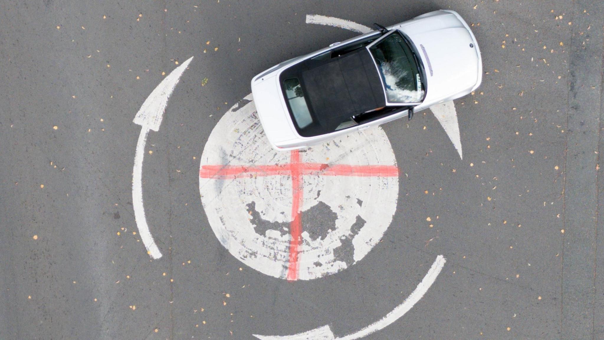 A white car drives over a mini-roundabout which has had a red cross painted on the white circle.