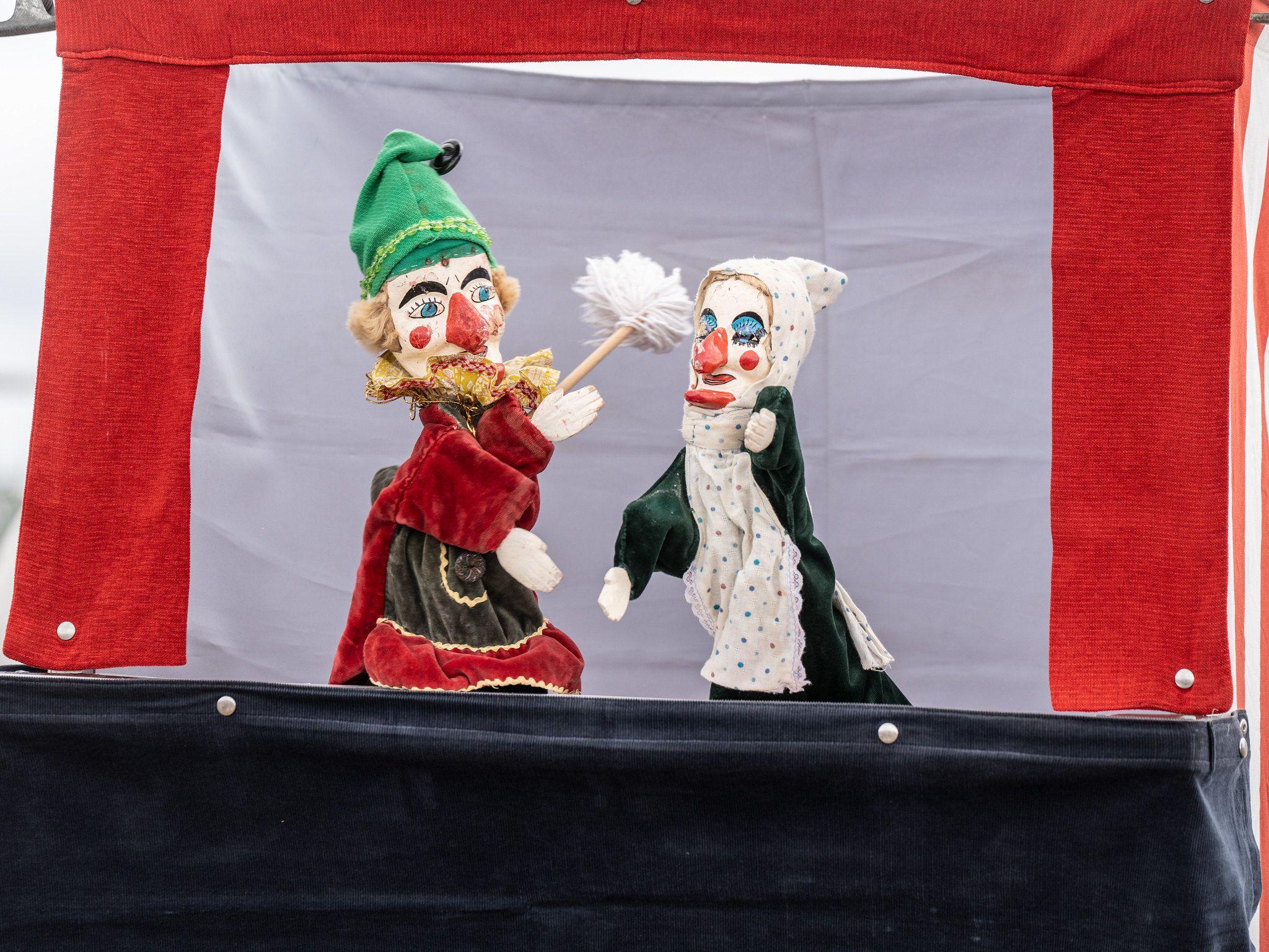 A traditional Punch and Judy show with puppets of the two framed by a red velvet stage. Punch is tickling Judy with a duster. 