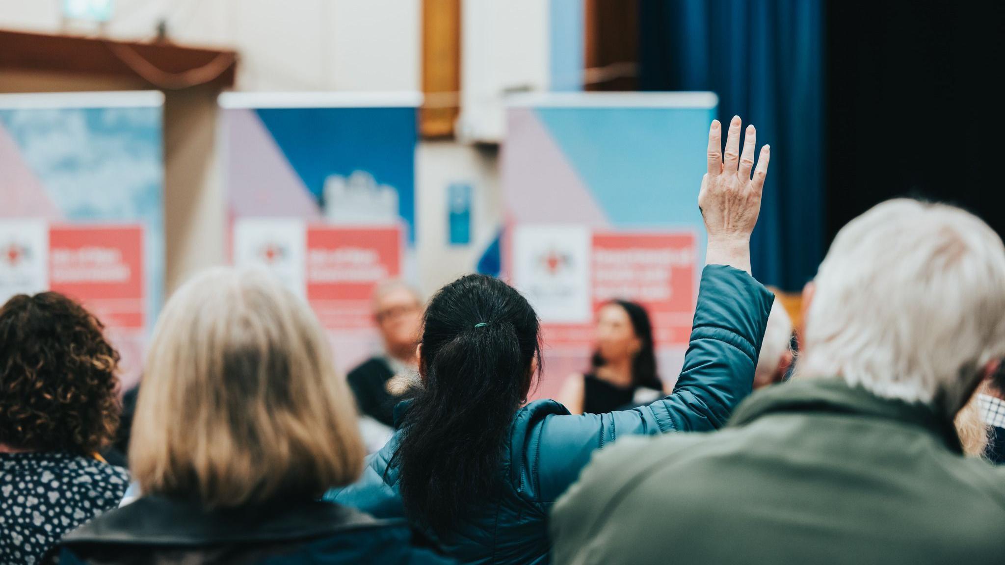 A woman wearing a blue coat raising her hand at a meeting, she faces ministers that have been blurred by the camera. She sits in between other people who are looking in the same direction.