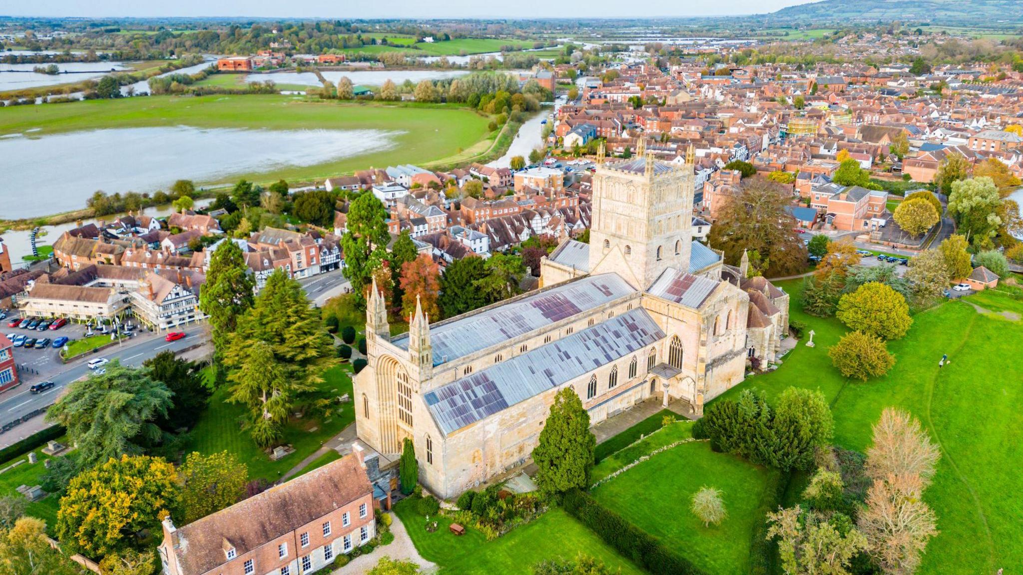 An aerial image of Tewksbury Abbey with the town and floodplains in the background. There is lots of lush green space and the roofs of the houses are terracotta tiles