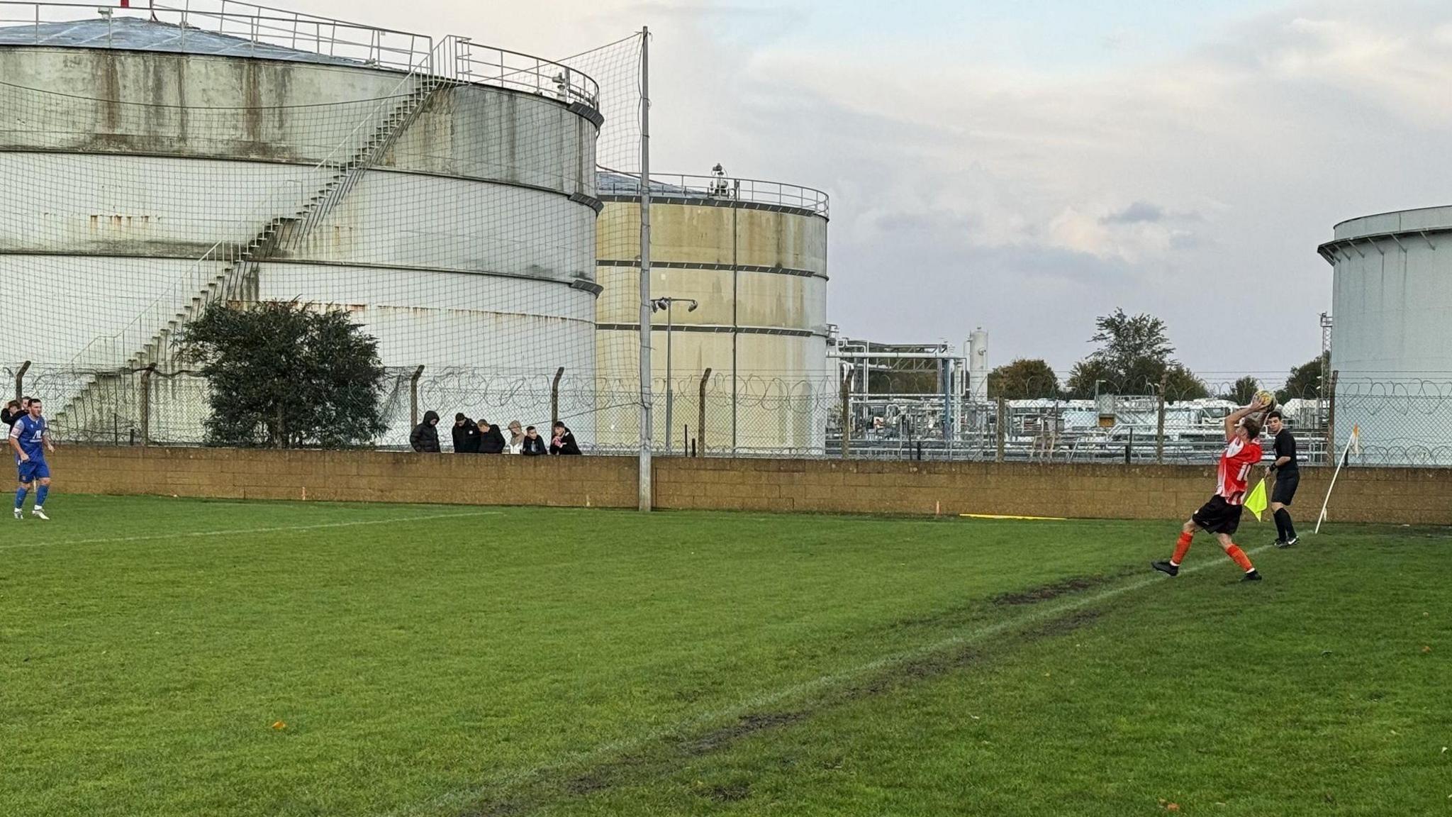 A player takes a throw in on the side of the pitch with oil storage towers visible in the background