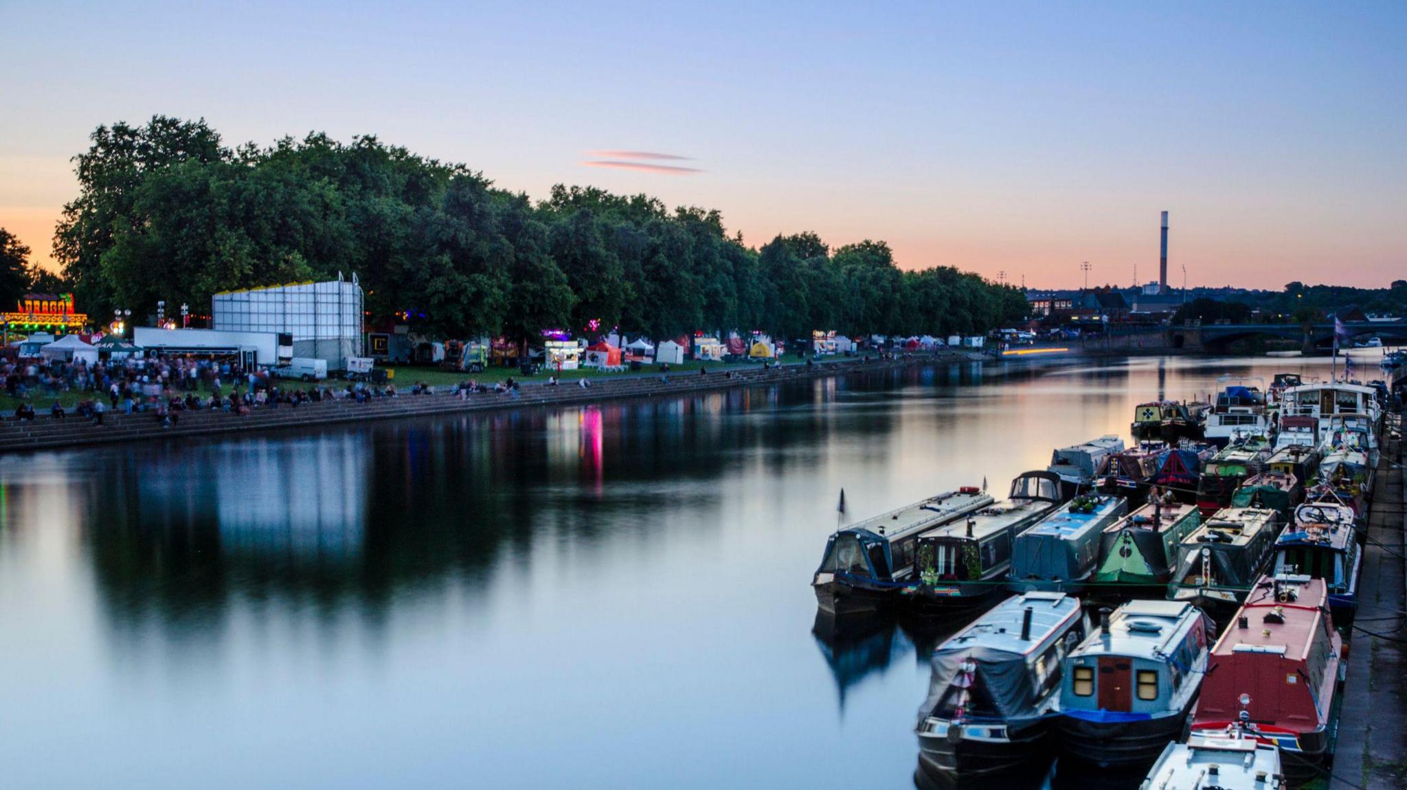 An image of the River Trent in Nottingham showing a number of moored long boats and a funfair on the other side of the river. People are sitting along the Victoria Embankment as the sun sets in the background, behind a row of trees.