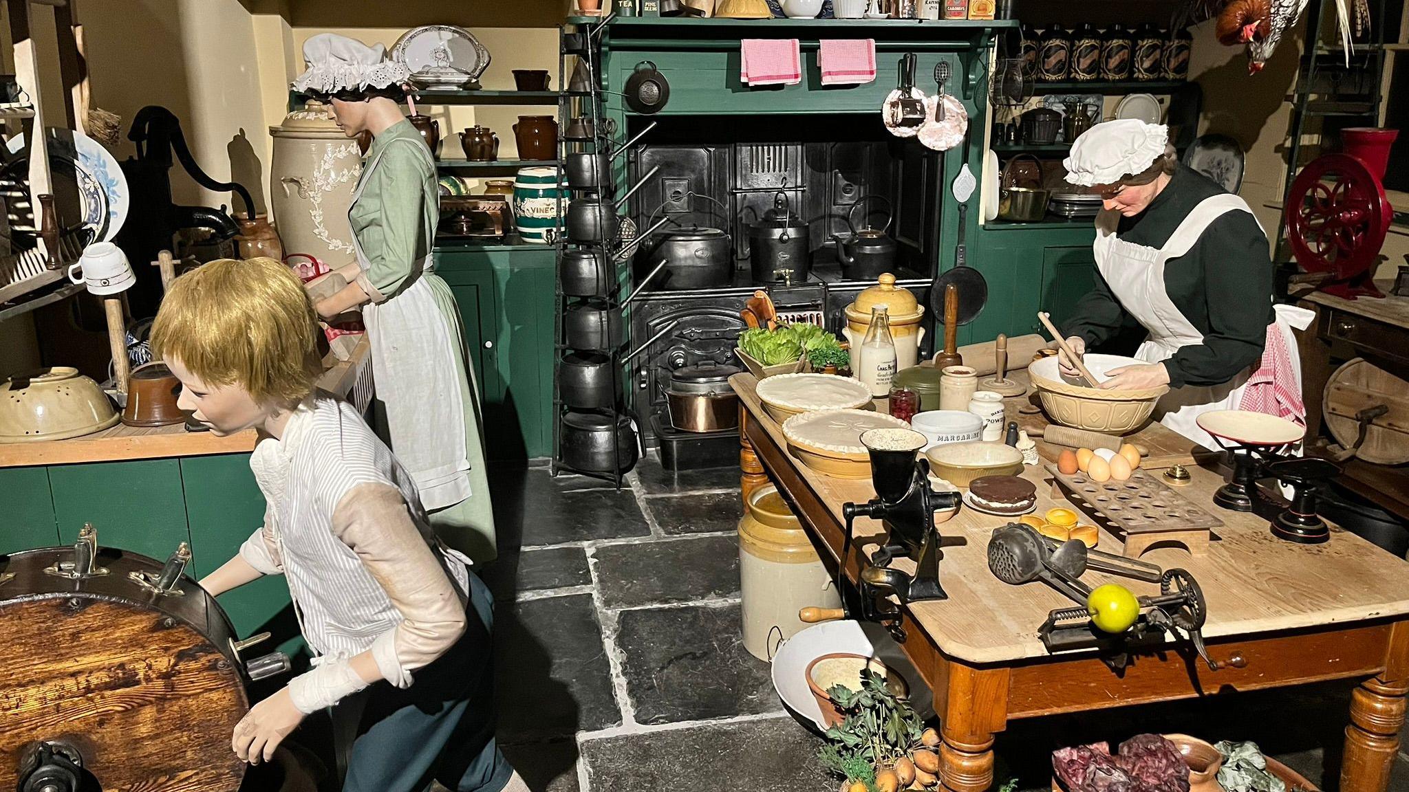 A scene depicting a Victorian kitchen, with a woman using a mixing bowl on a wooden table, another woman standing beside a counter, and a child also in the kitchen