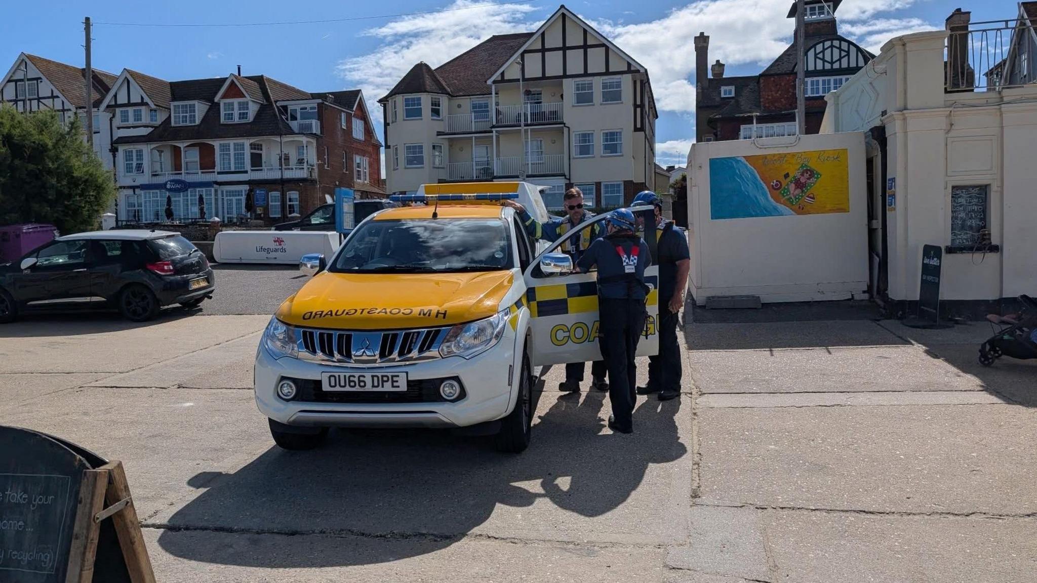Three members of HM Coastguard stand by a yellow and white marked vehicle on Westgate beach on a sunny day. Behind them are several seafront buildings and two more parked cars.
