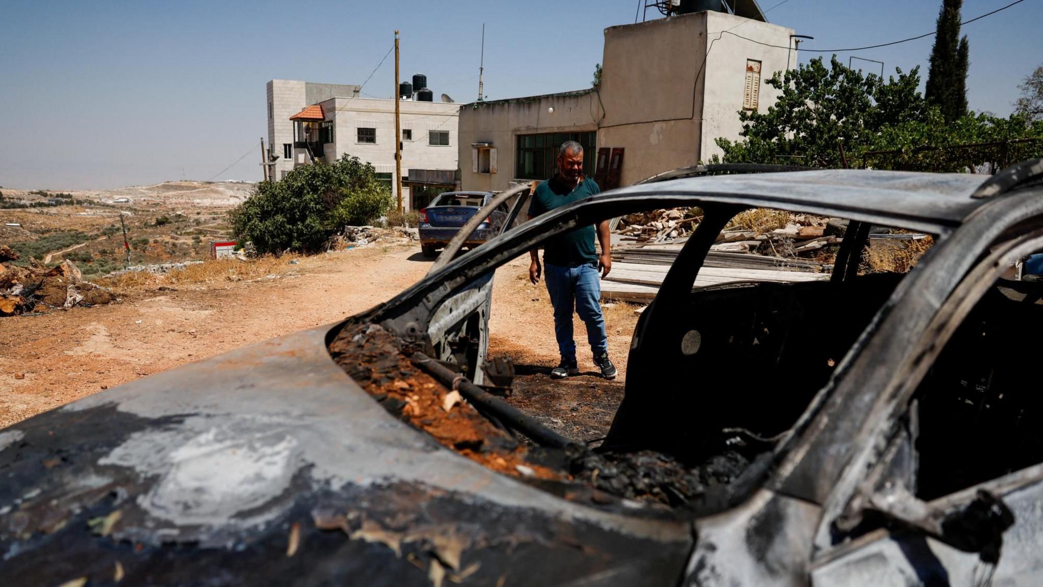 A Palestinian man stands next to a burnt car after an attack by Israeli settlers in Kafr Malik, in the Israeli-occupied West Bank (26 June 2025)