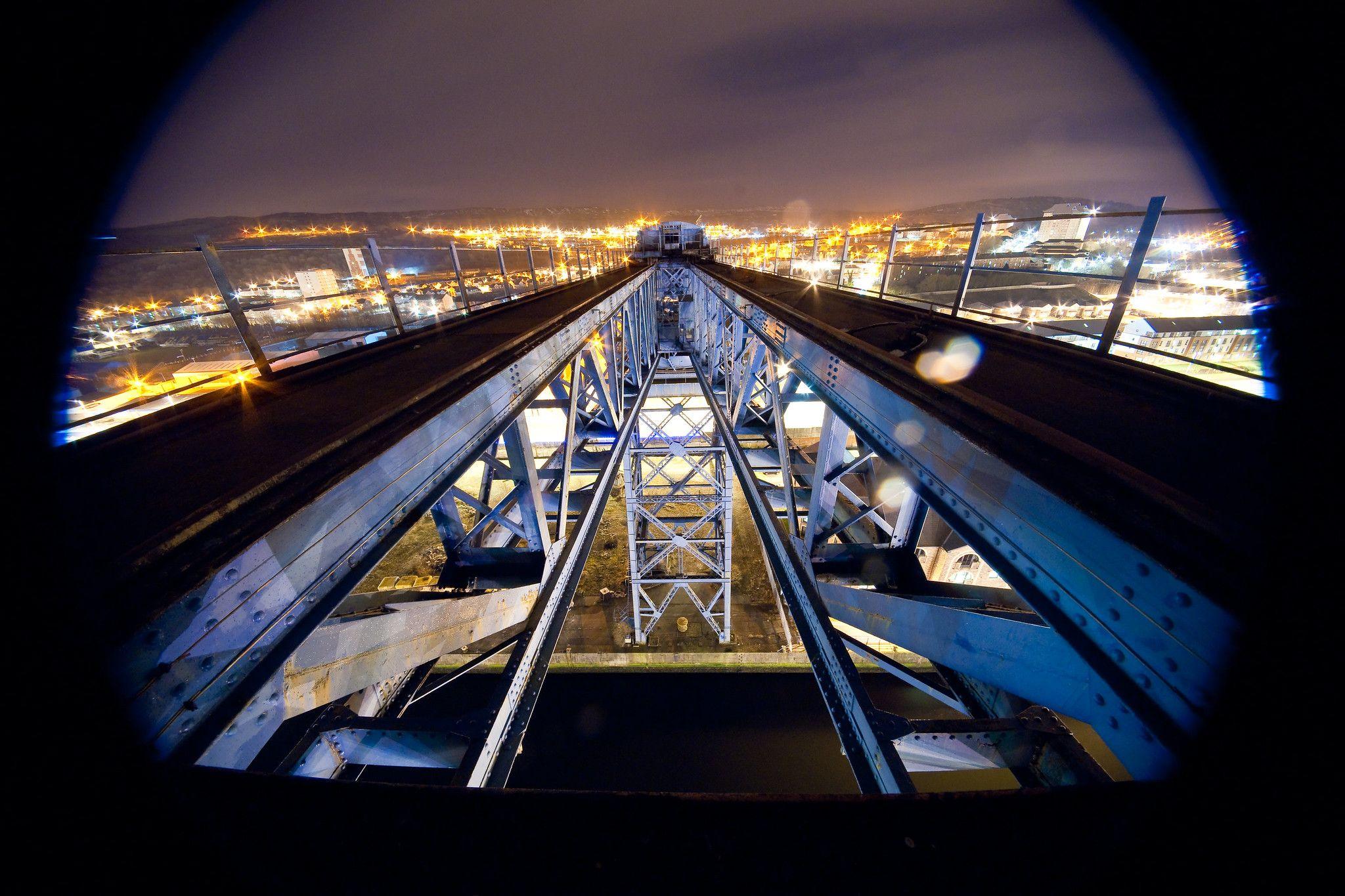 A blue painted crane photographed at night. The crane is constructed of giant latticed girders. There are orange and white lights in the distance.