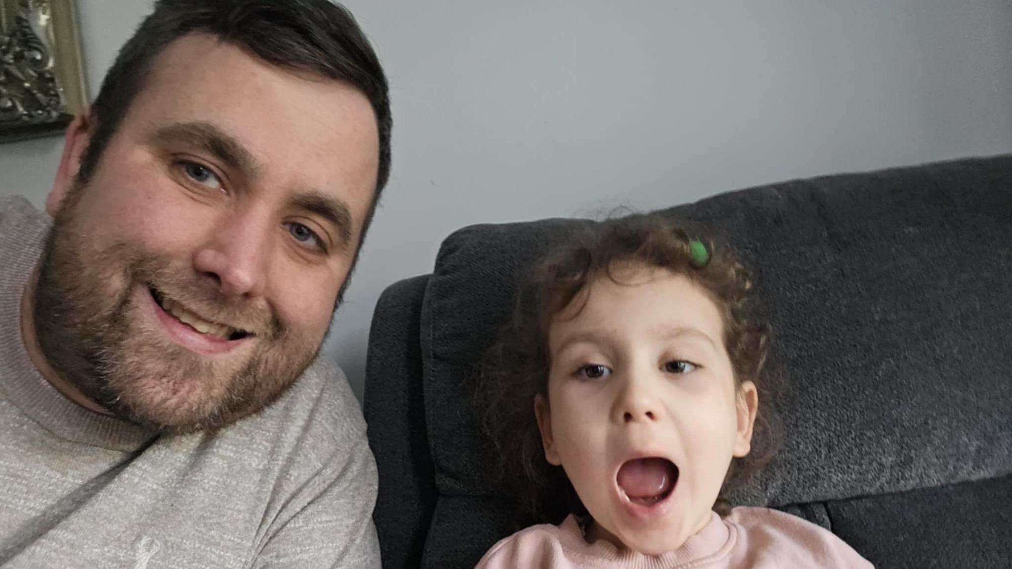 Columba and Maisie together on a grey sofa, Columba is smiling and wearing a grey sweatshirt and Maisie has her mouth open wide while wearing a pink sweater