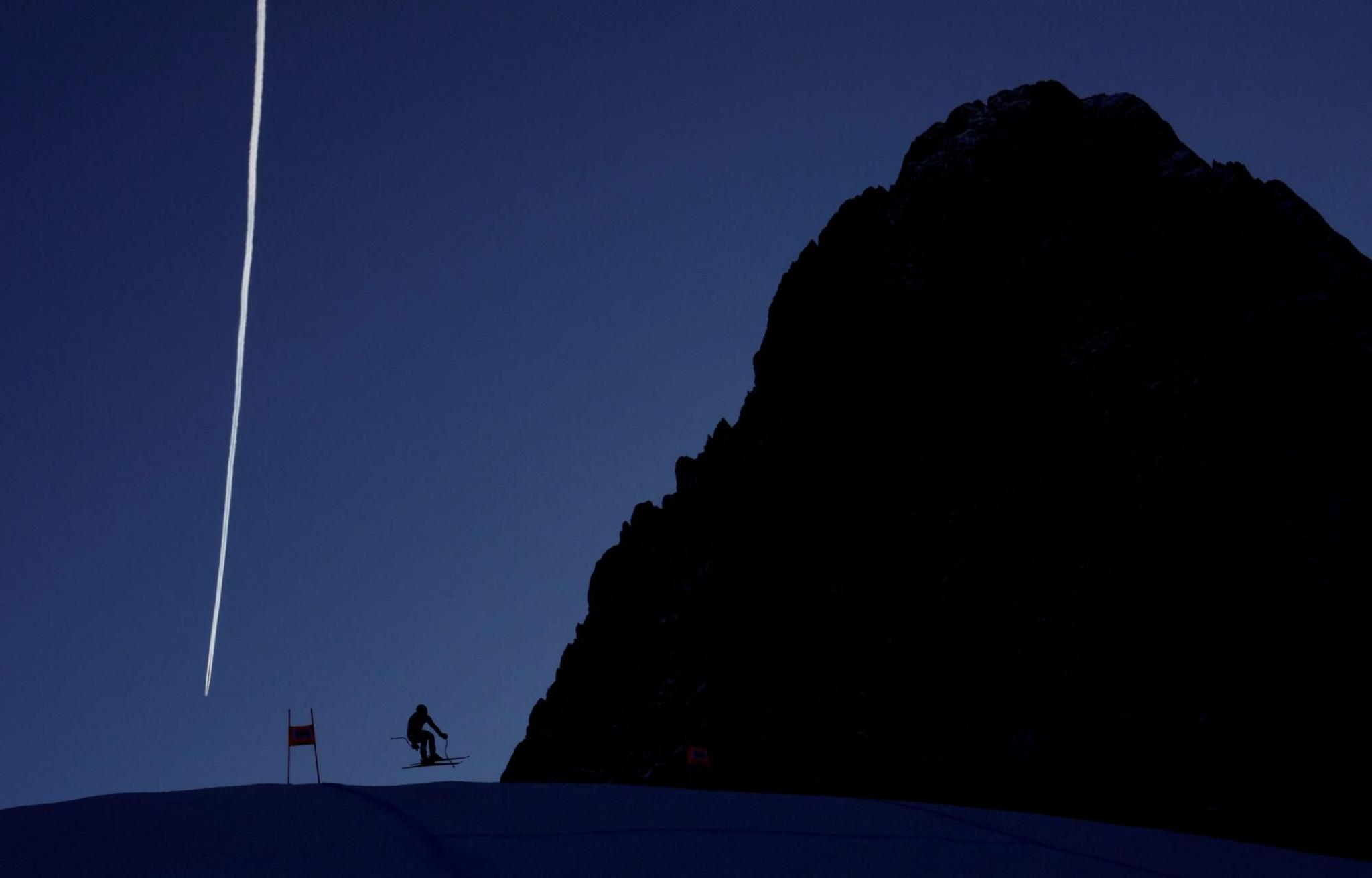 Skier Kyle Negomir is silhouetted against the landscape and blue sky as he practises a ski run at Val Gardena, in Italy. He appears tiny beside a giant rock, which is also in silhouette, while a white vapour trail stands in the sky behind them both. 