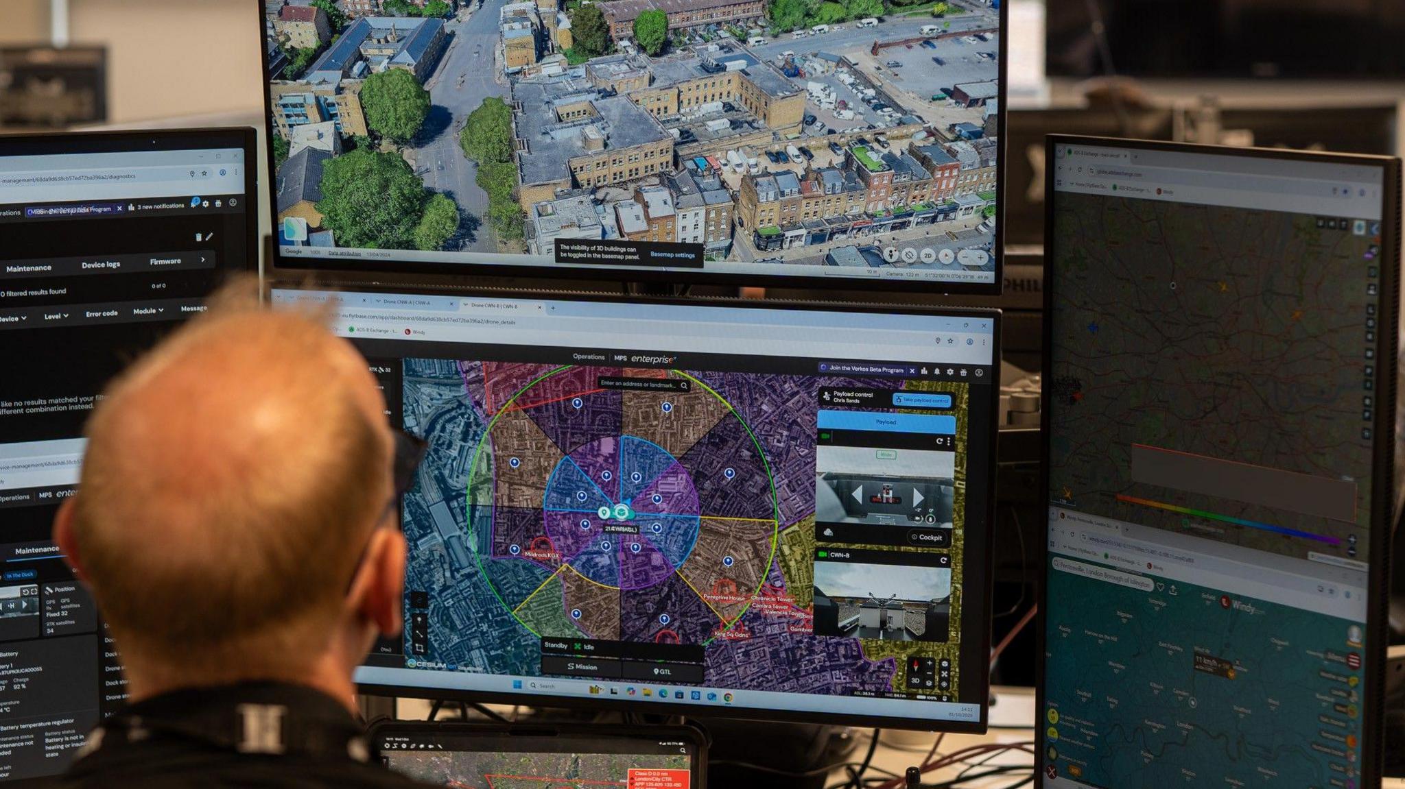 A police officer sits in front of a bank of computer monitors with his back to the camera. One monitor has a drone image of a street scene.