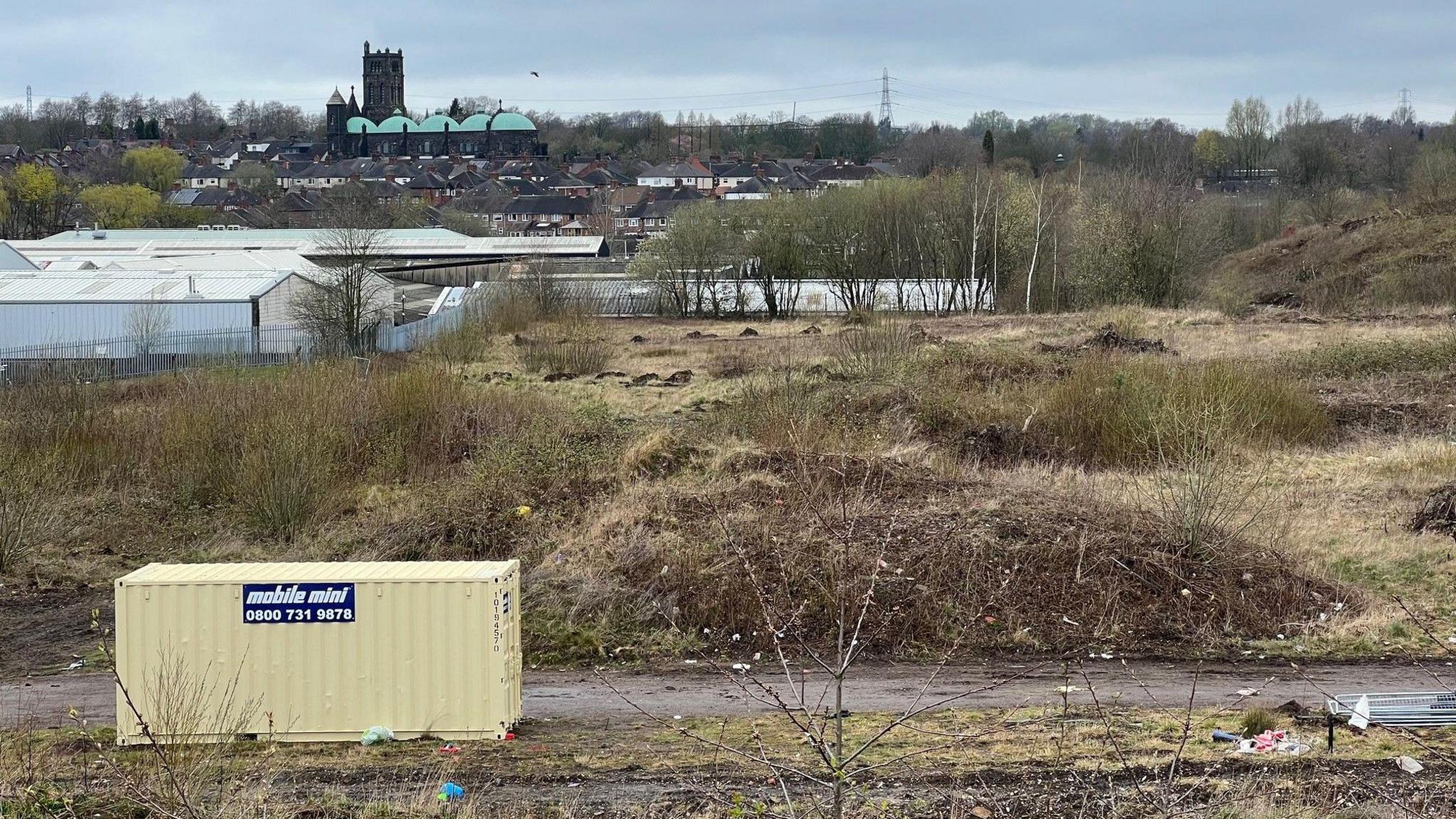 A shipping container in the foreground, sat in a patch of scrubland, with a industrial units and a town centre in the distance.