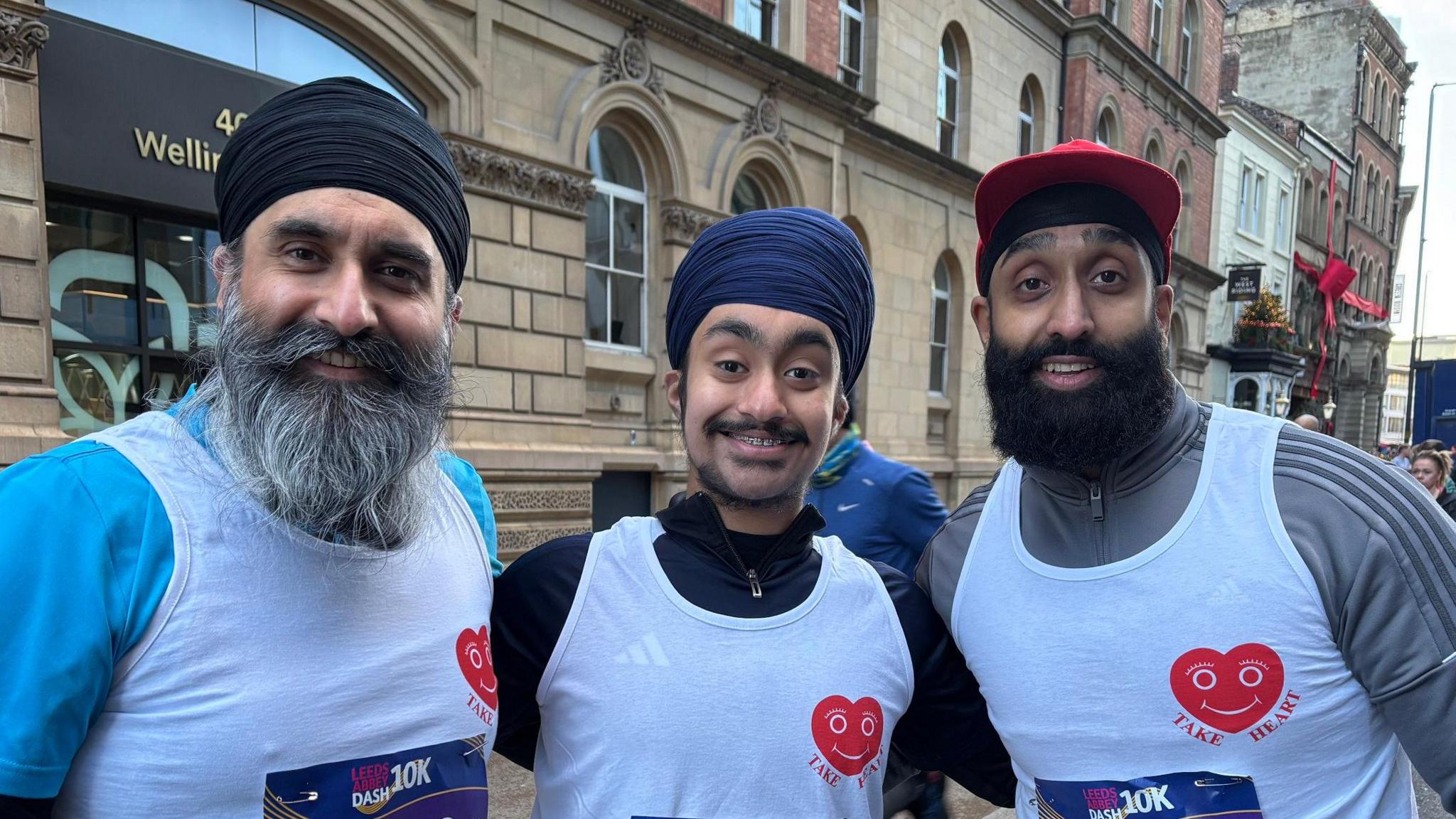 Three men wearing white running vests with heart logos and race bibs posing together on a city street during a 10K event. They are dressed in athletic gear, including long-sleeve tops and head coverings.