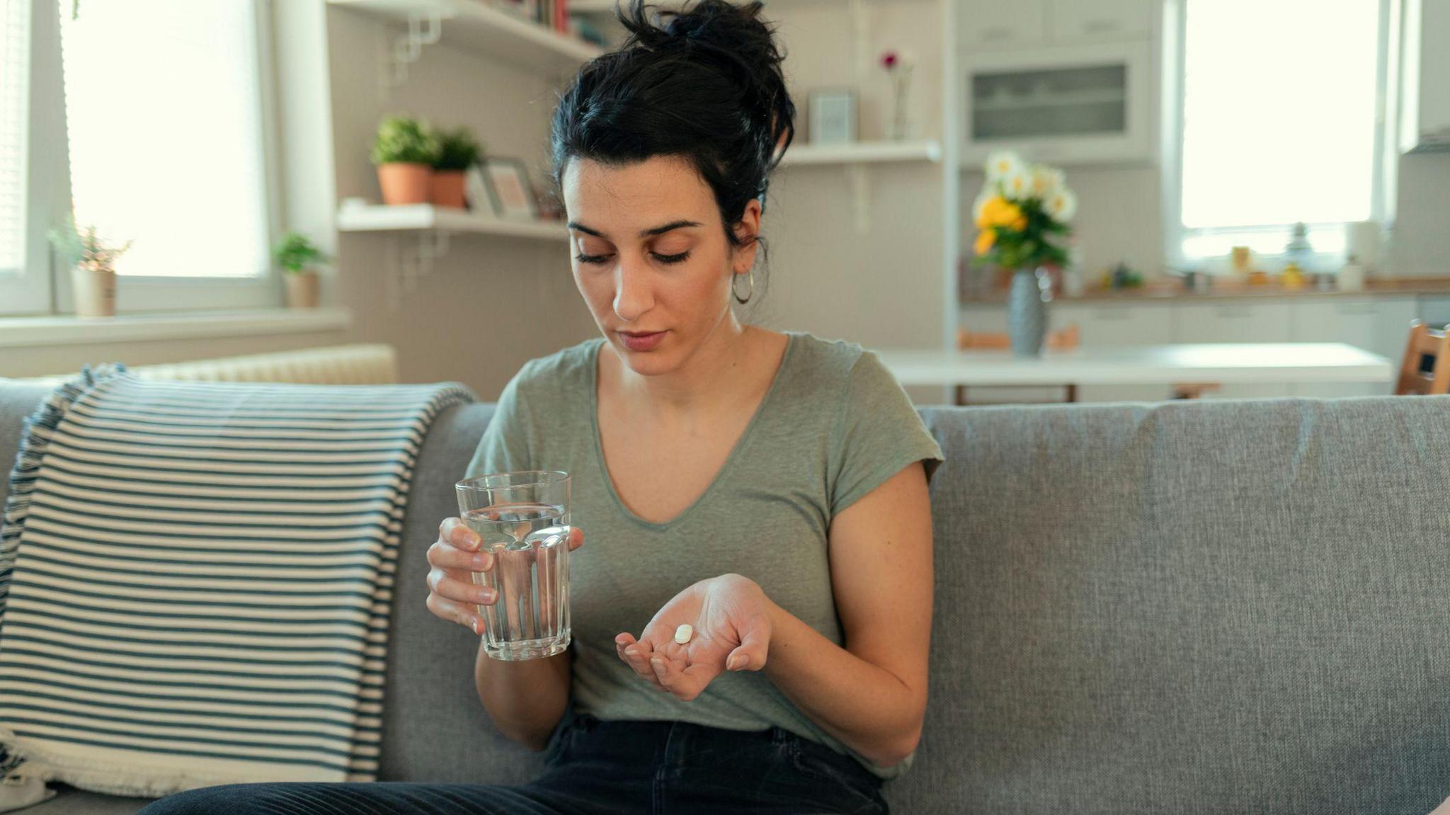 Young woman is sat with a glass of water in one hand and a white tablet in the open palm of the other. She is looking down so you can't see her eyes and her hair is tied up. She is sat on a grey sofa in what looks like an open plan living room with the kitchen behind her.
