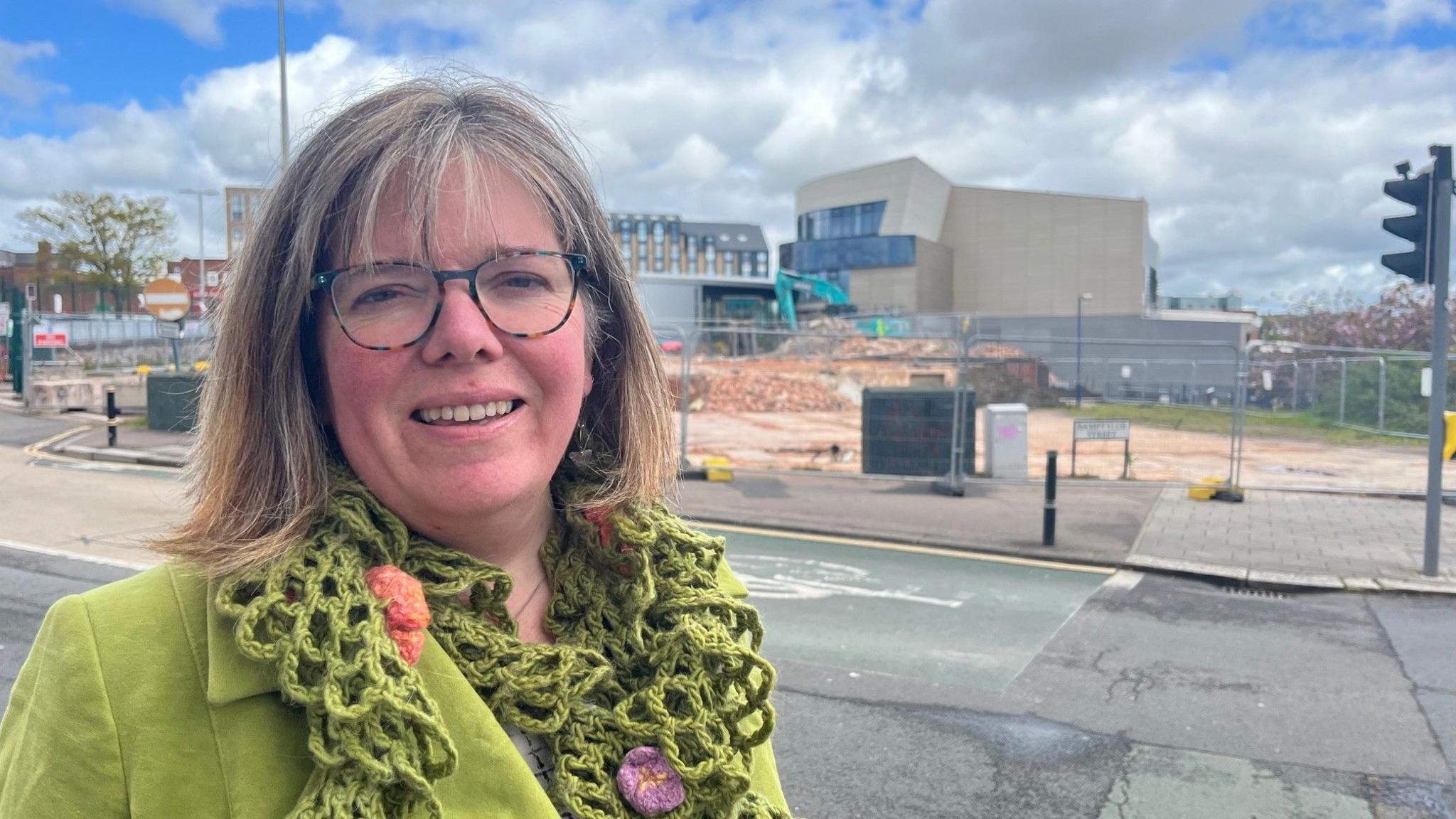 Diana Moore, Exeter Green Party leader, is wearing a green jacket and scarf and glasses. She is standing in front of the old bus station site when it was demolished in April 2024