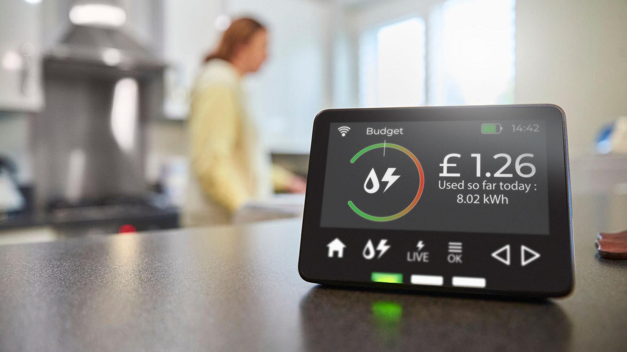 A smart meter energy display on a kitchen surface with the screen saying £1.26 of energy has been used today. A woman in the kitchen is blurred in the background.