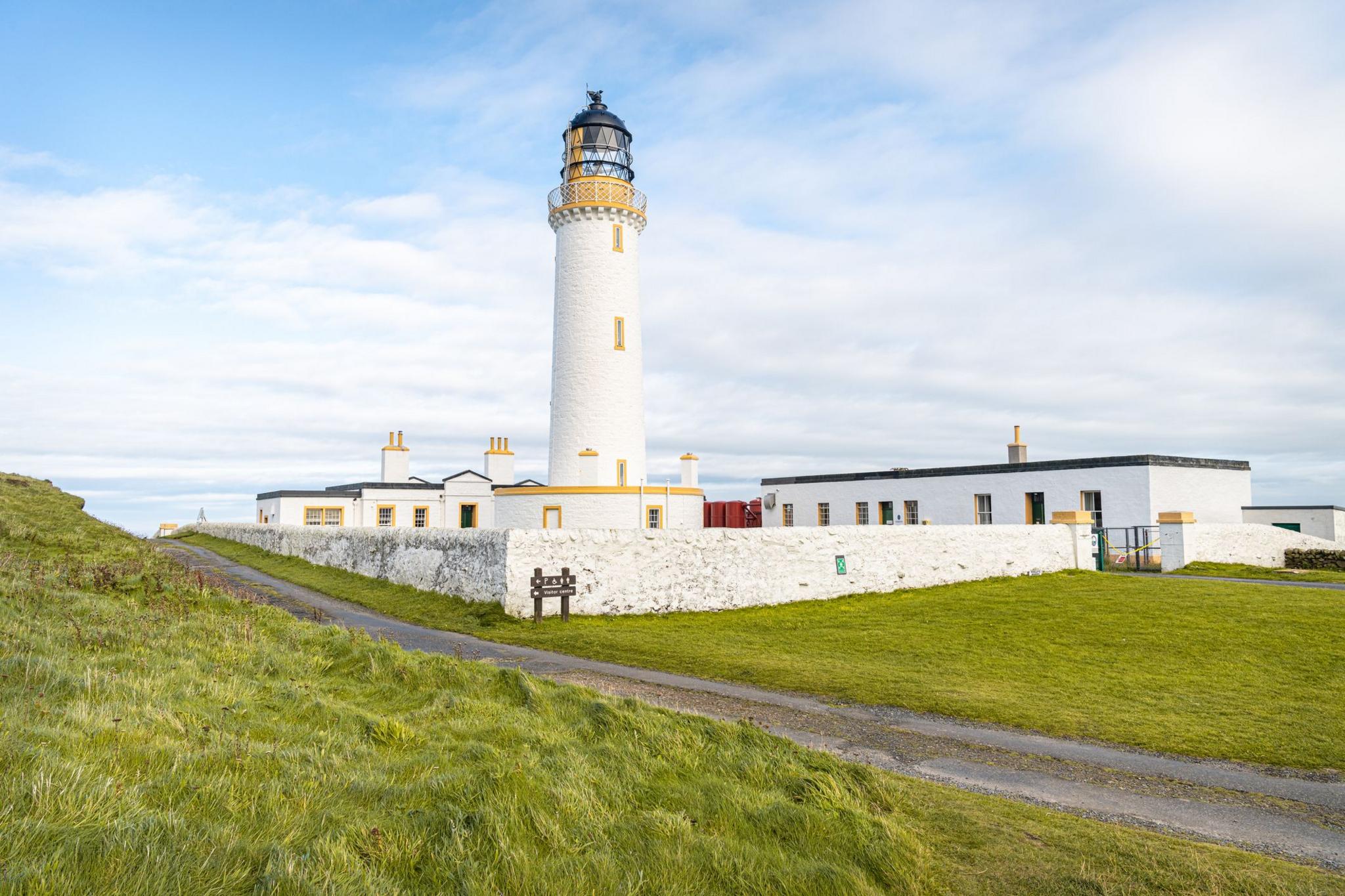 A white lighthouse building with yellow windows sits at the end of a pathway through grassland