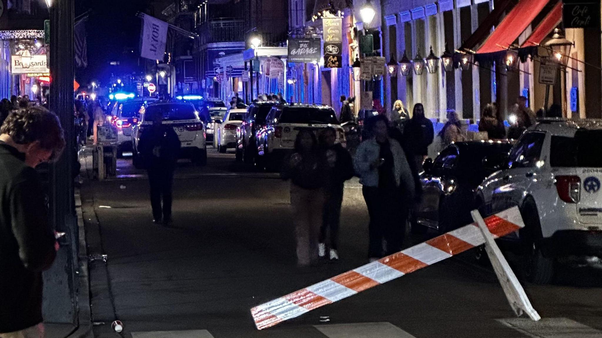 Crowds on a busy high street with police cars in the background. A red and white striped barrier lies half on the ground with the other end supported by a white stand. People mill in between cars on the road.