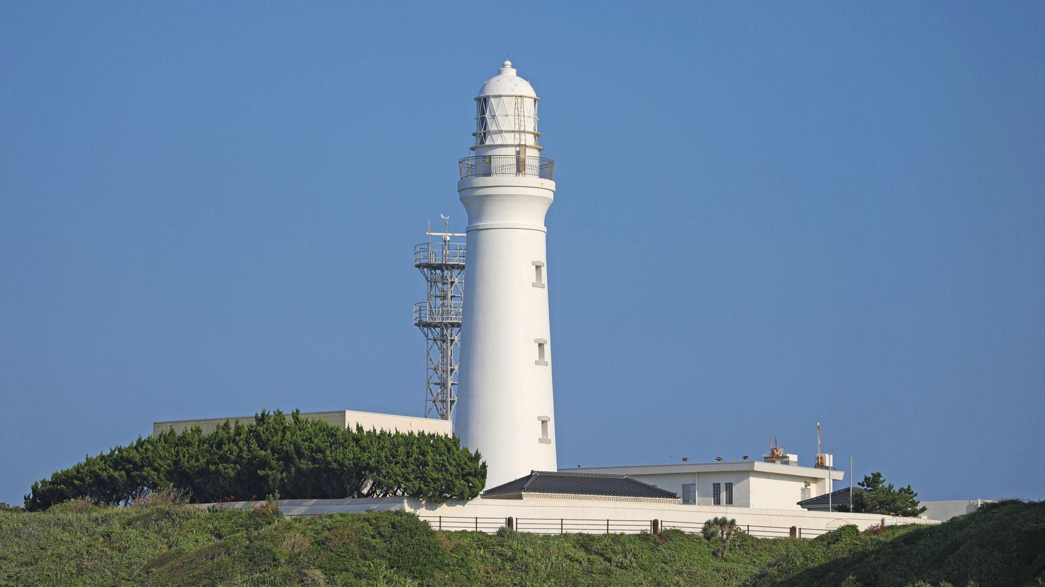 A white lighthouse surrounded by other low buildings and trees juts out into a blue sky