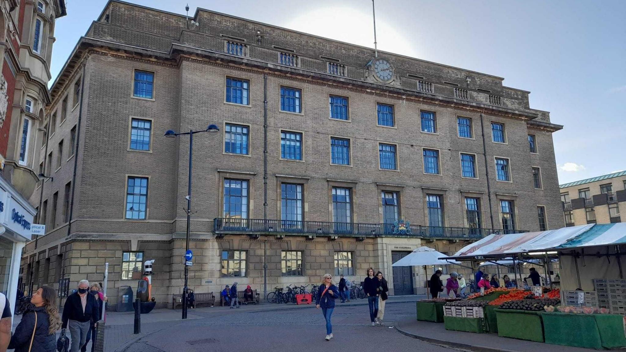 Exterior of the Guildhall in Cambridge. A large square brick building with large windows and a clock at the top. There are market stalls selling fruit and vegetables opposite and people mill about.