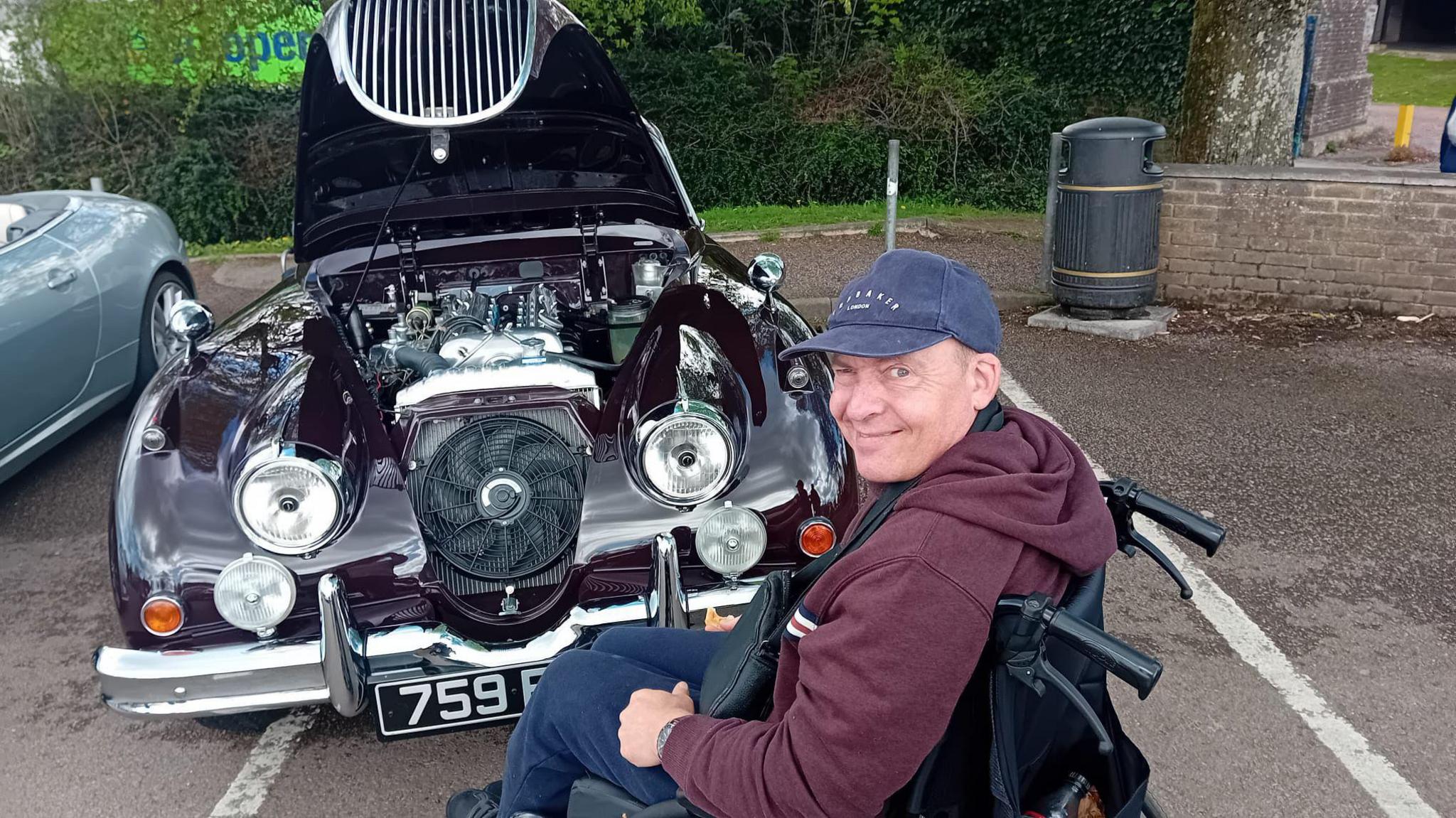 Richard Rudd is seen next to a vintage car. The bonnet is open and the engine is visible. Mr Rudd is sitting turned away from the camera in his wheelchair, and he has his head turned to the person taking the picture and looking happy. The car is in a car park, and there is a hedge and a wall in the background.