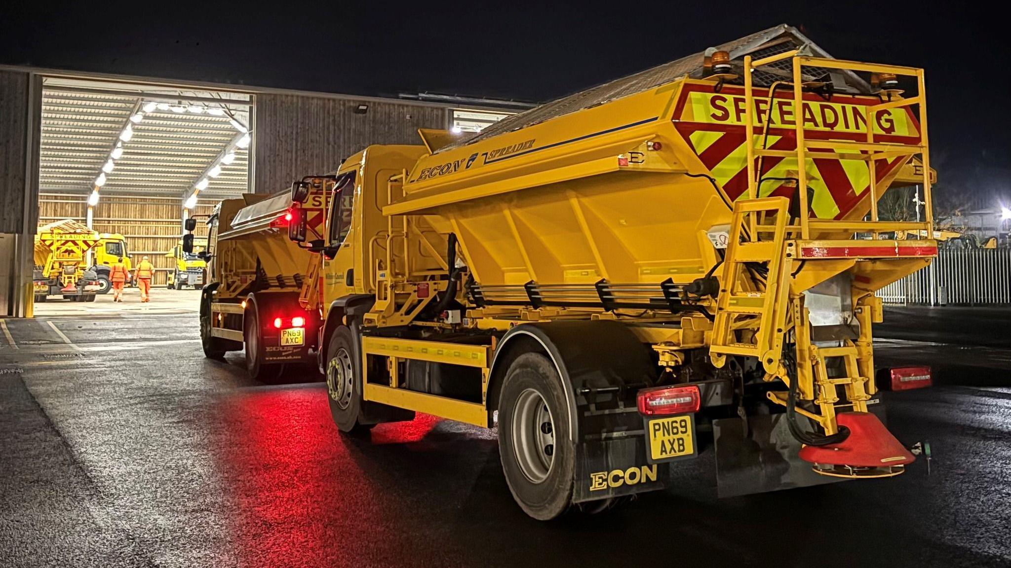 Two large yellow gritter trucks with red and yellow hi-vis panelling at the back. They are reversing out of a large industrial warehouse, which is lit up with white lights. Outside it's dark and there are some road workers walking in the warehouse. 