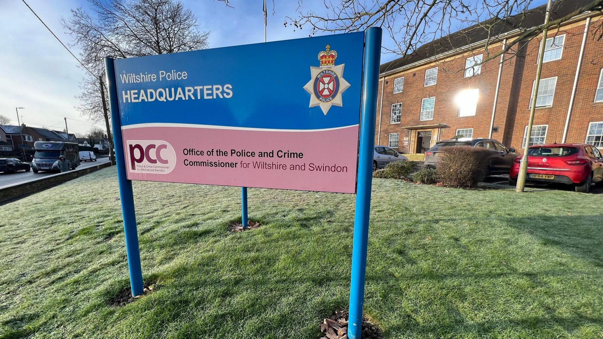 Wiltshire Police Headquarters sign, which is blue and yellow, on a lawn in front of the building. There are some cars parked in front of the building.