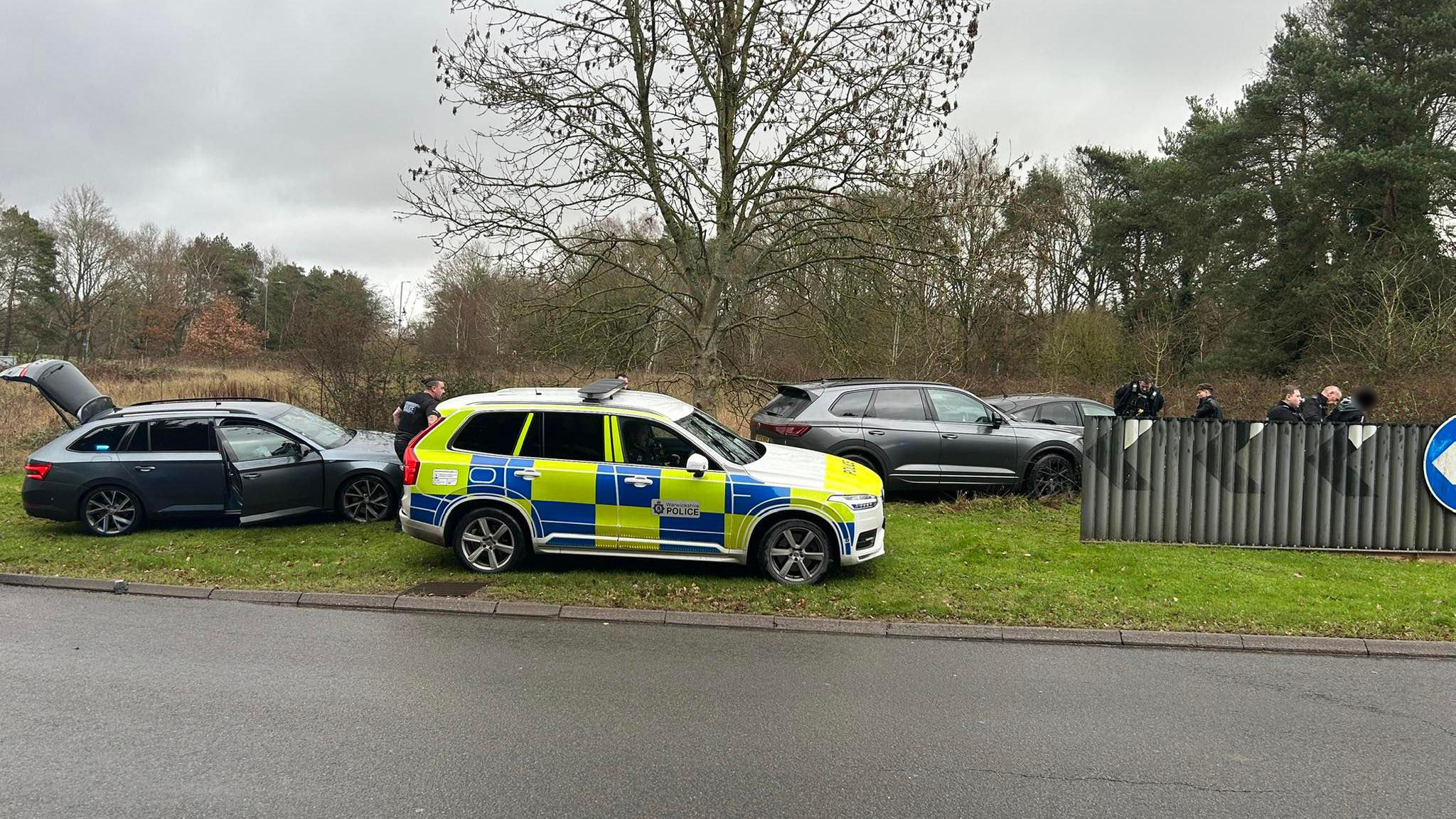 A yellow and blue police vehicle sits on a grassy bank beside the A46 in Warwickshire. Three other black vehicles are parked nearby. Police officers can be seen behind a road barrier. 