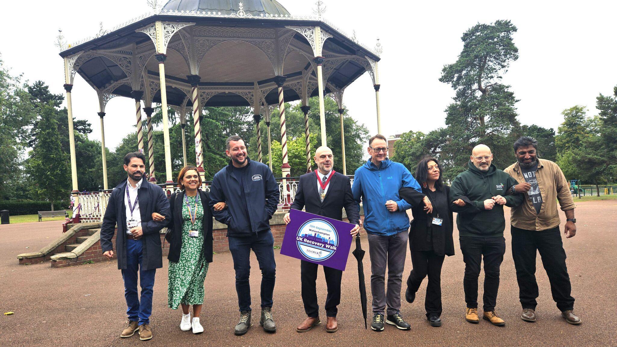 Eight people, six men and one woman are standing in a line in front of a bandstand, linking arms. A man in the middle, the city's mayor, is holding a purple card that says "UK recovery walk"