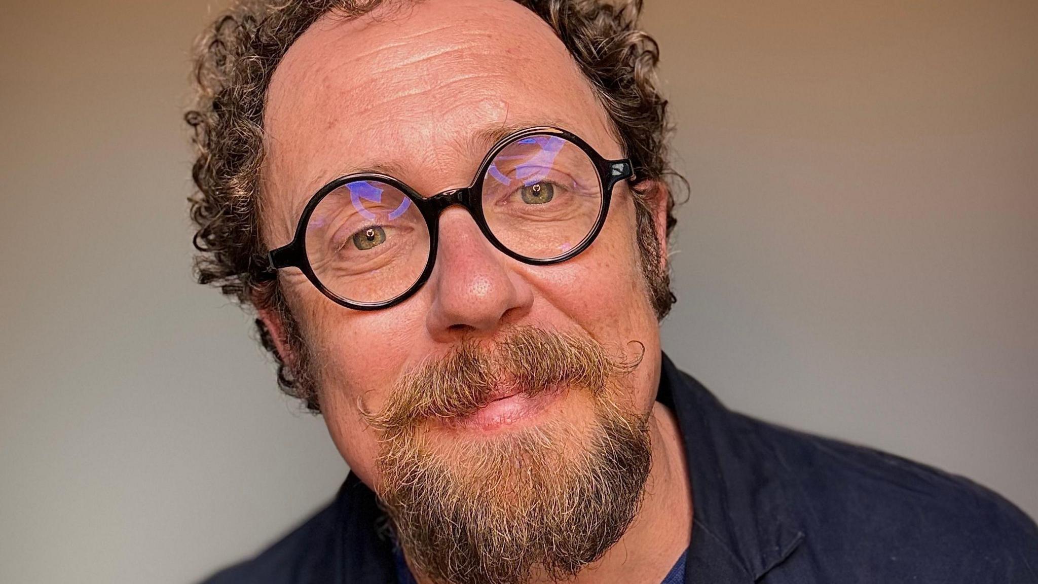 Bob Golding has brown curly hair and a moustache and beard, he wears black-rimmed round glasses and is looking straight at the camera.