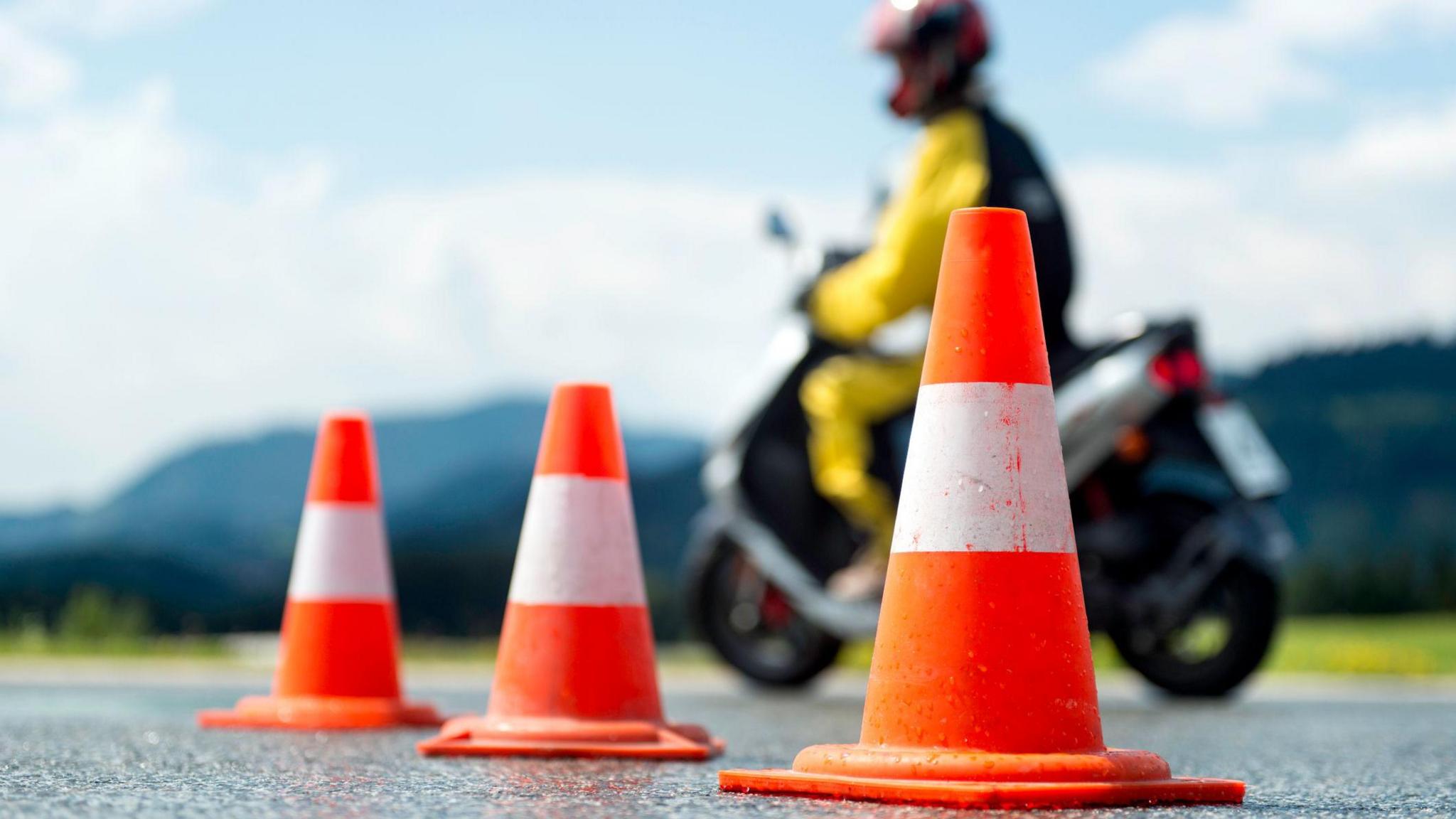 Cones on the ground with a person driving a moped in the background.