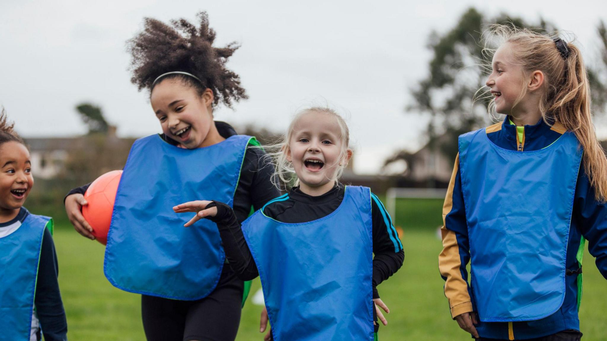 A group of children smile as they walk wearing bibs and carrying a football.