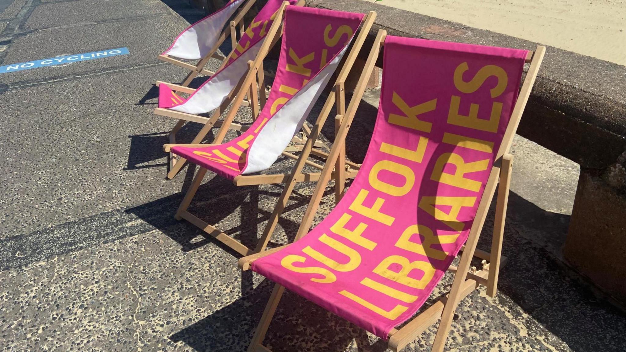 Pink deckchairs with Suffolk Libraries written in yellow print on a path next to Lowestoft beach