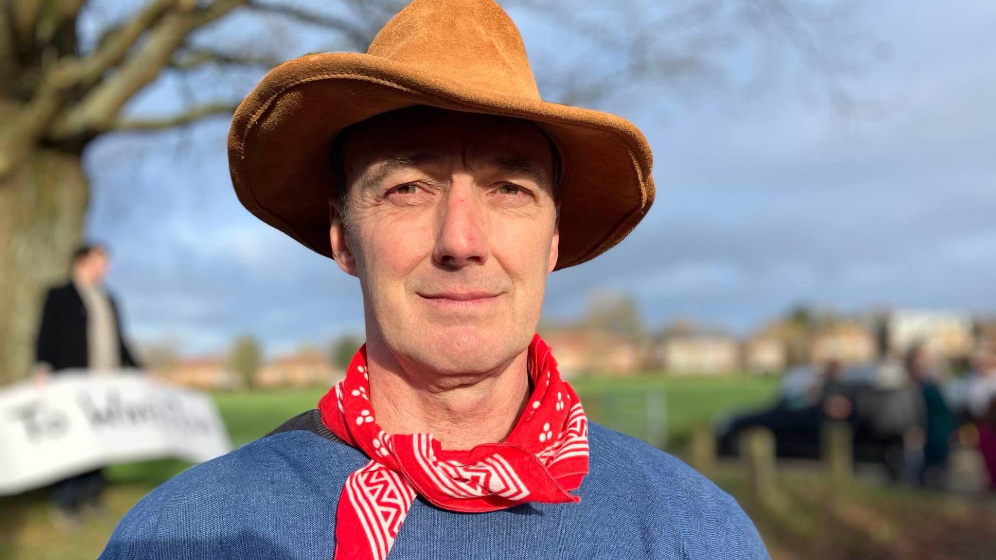 A man looking at the camera wearing a brown cowboy-style hat, red and white neckerchief and blue jumper. Behind him is a blurred tree and field