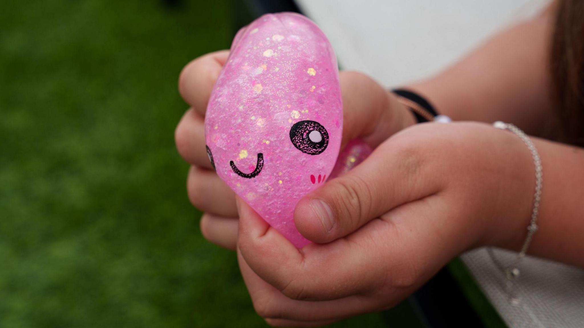 Child’s hands squeezing a pink glitter squishy dumpling toy. The toy has a smiley face on it. The child is wearing a silver bracelet.