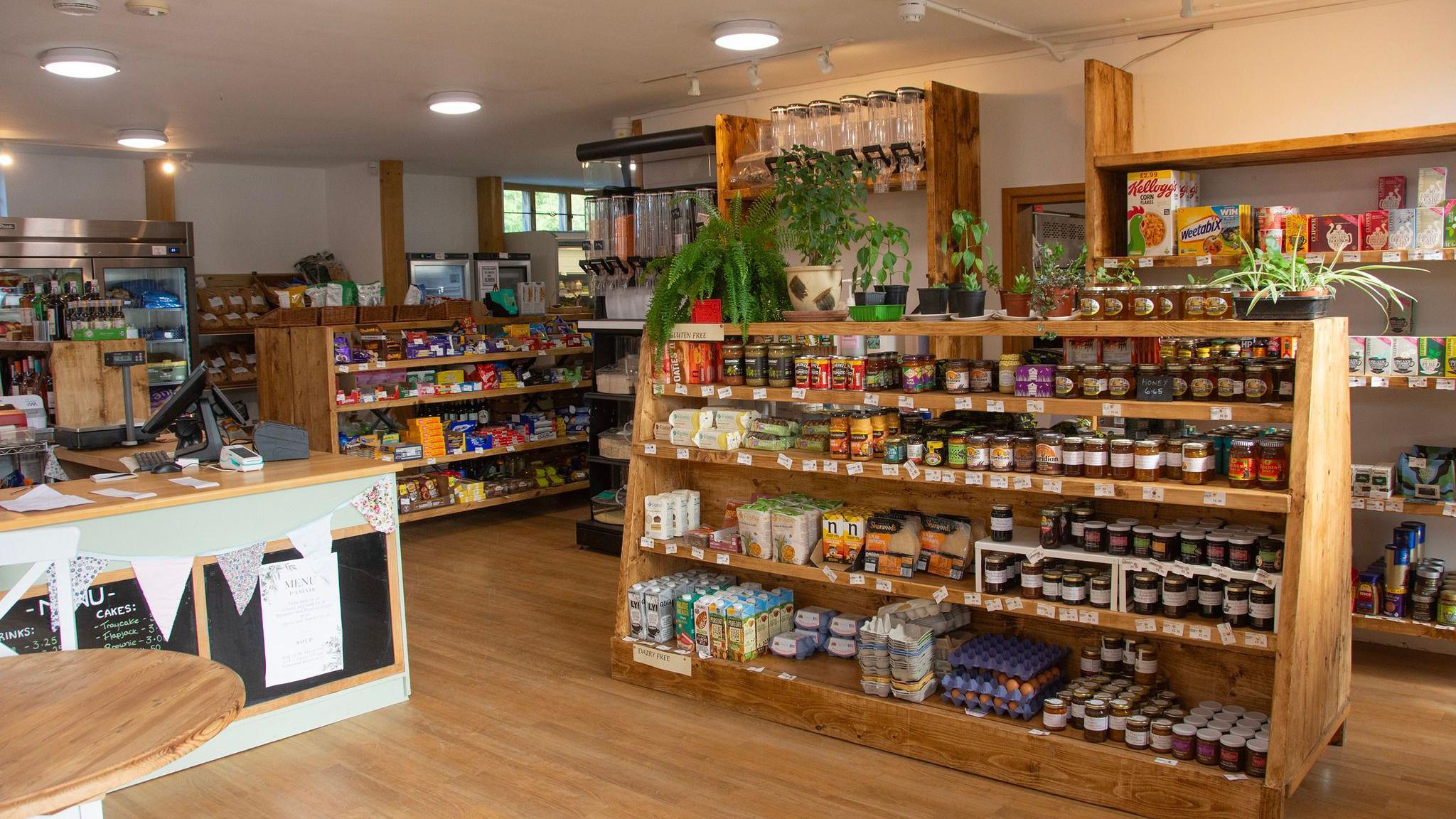 The interior of the newly restored café. There is a warm orange glow from the sun outside. In the centre of the room there is a large wooden shelving unit which has jars of preserves, flower, sugar and lactose free milk stacked neatly. On the top are potted plants. To the left of the image is the till area with fabric bunting hanging from the counter. In the background there are refillable jars and more wooden stands full of products.