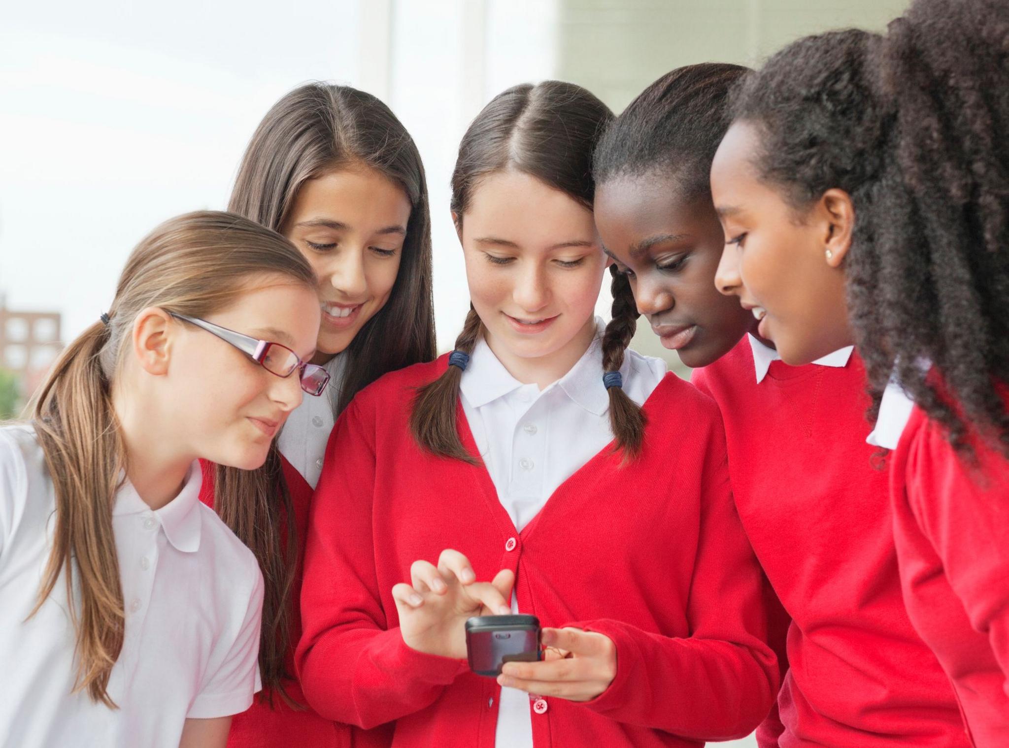 a group of pupils in school uniform look at a mobile phone held by one of the pupils in the centre as they all huddle round.