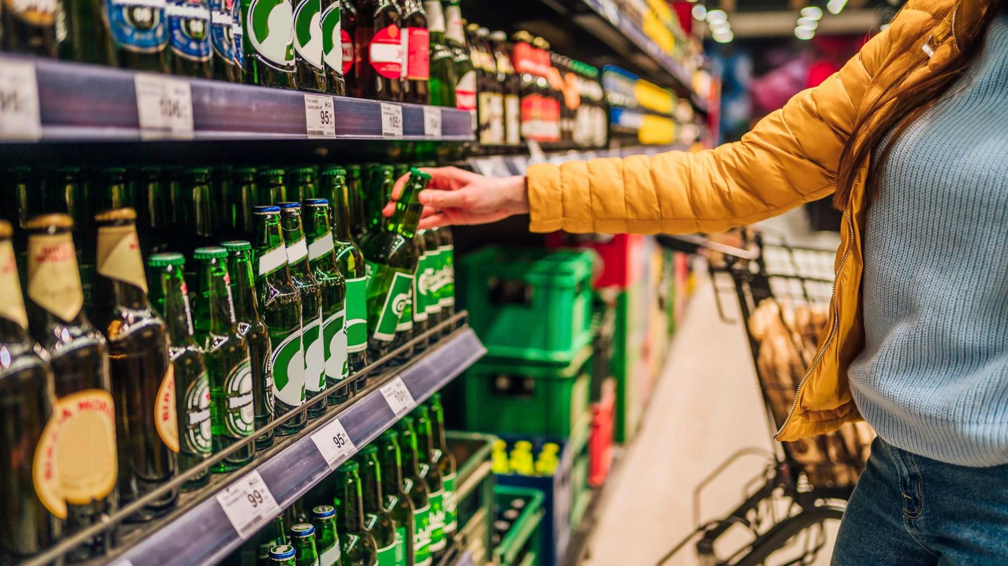 Rows of bottles of alcohol, woman in yellow jacket with blue jumper stretching out arm