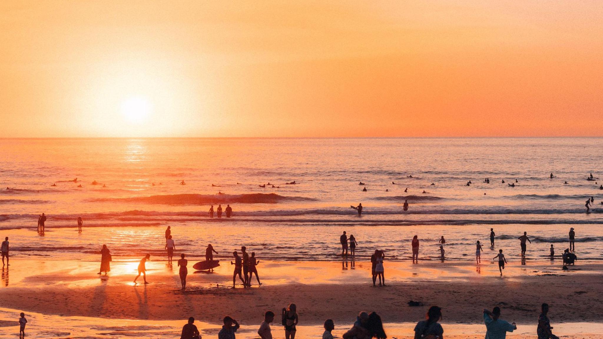 Crowds are walking in the sea holding surfboards as waves crash around them. The sun is golden and setting in the distance. 