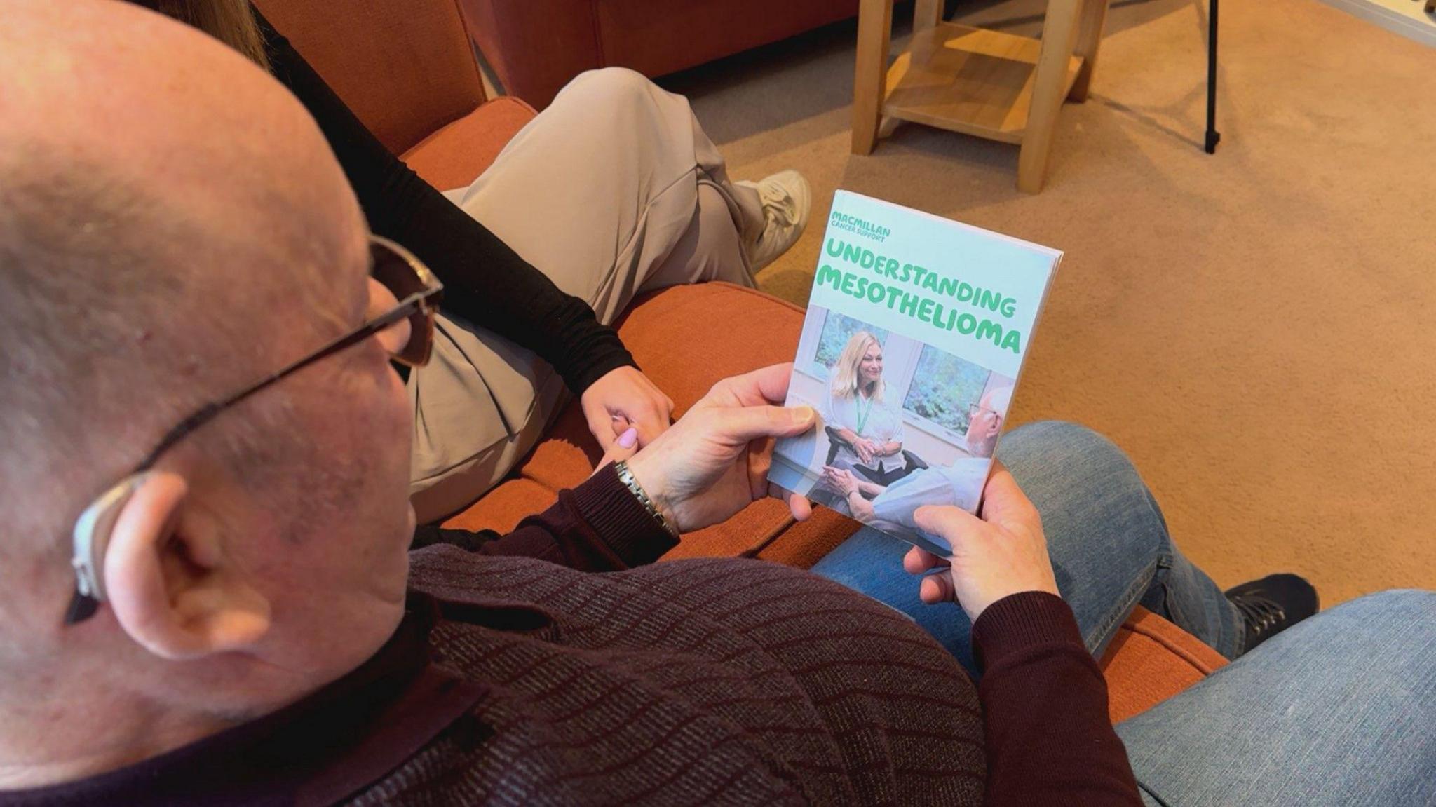 An over-the-shoulder shot of a man wearing jeans and a jumper holding a Macmillan Cancer Support guide to understanding mesothelioma.