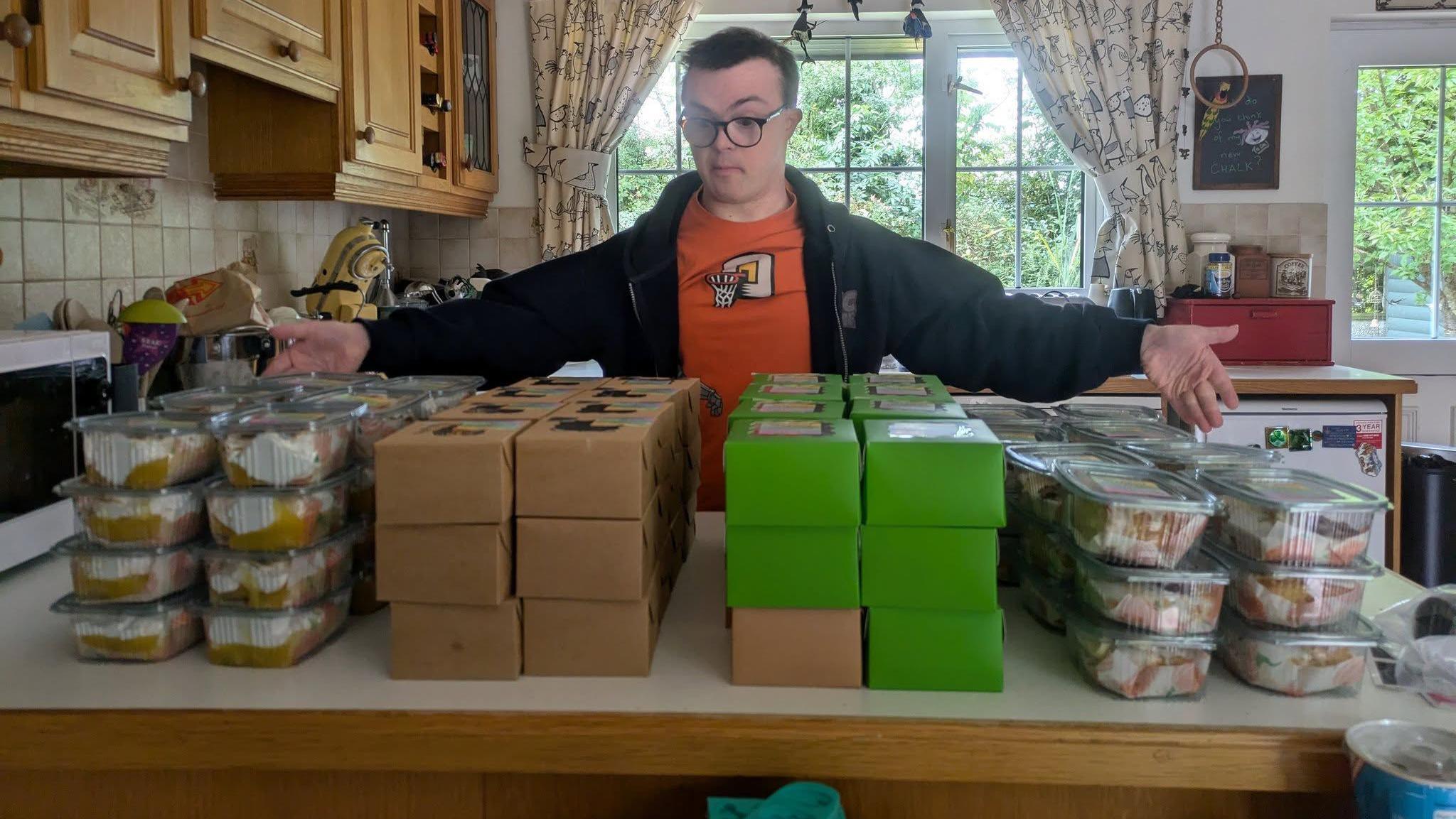 A young man with glasses is wearing an orange t-shirt and black jacket. He is standing looking at boxes of cakes and buns with his arms spread out to show the scale of the operation. He is standing in a kitchen. There is a glass double door and white curtains in the background. 