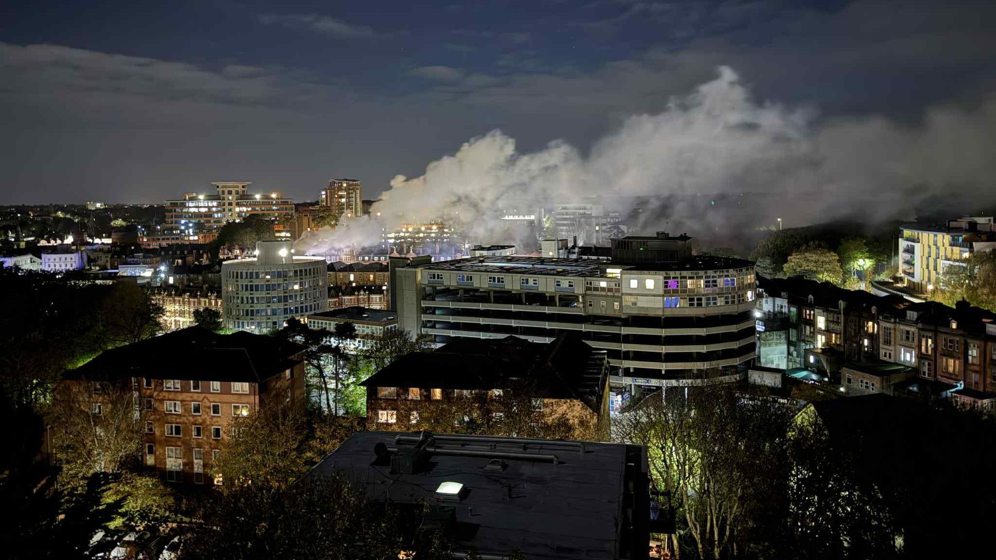 Plumes of white smoke billowing from left to right in Bournemouth Town Centre.  Lights can be seen in multiple buildings.  It is night.