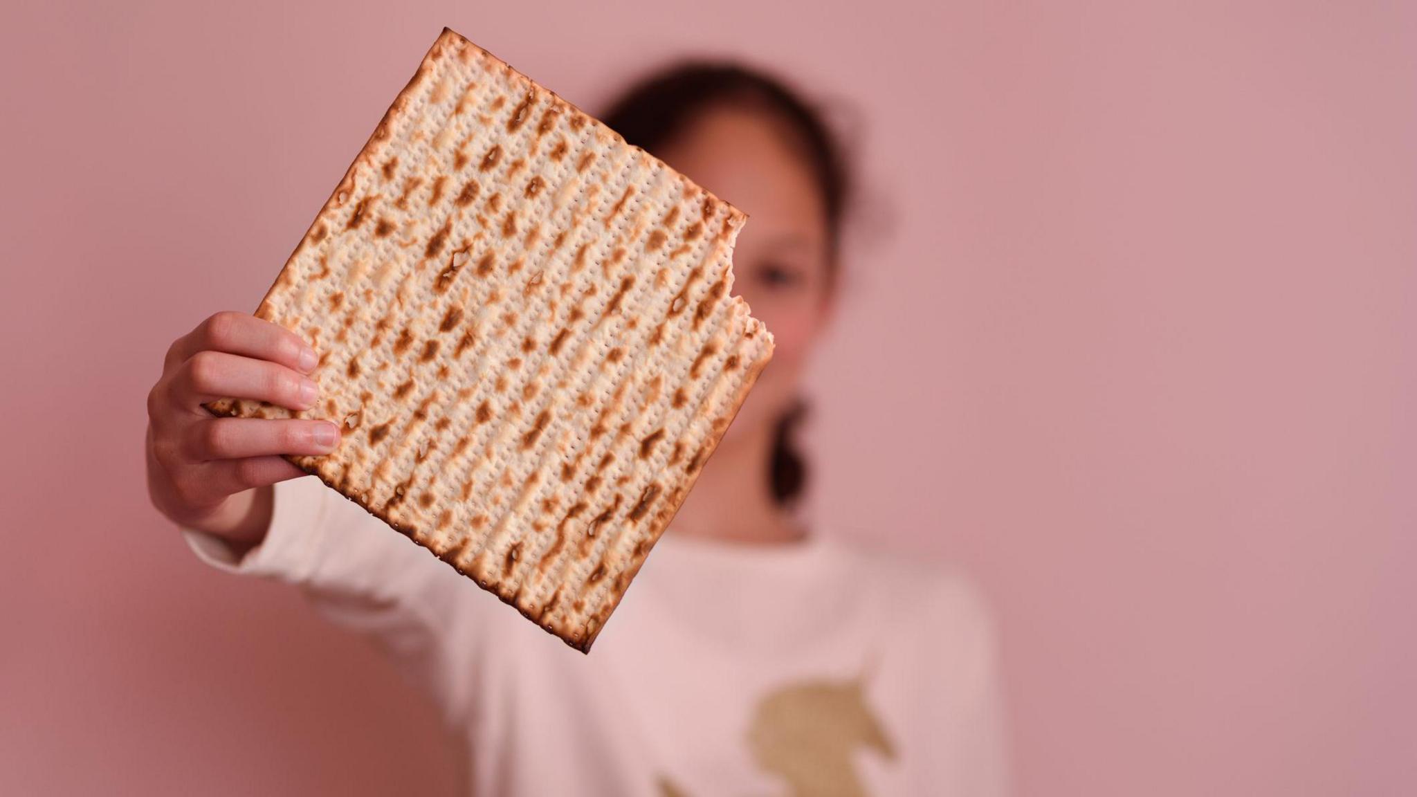 A girl who is blurred by the camera holds a matzah with a piece bitten off to the camera.
