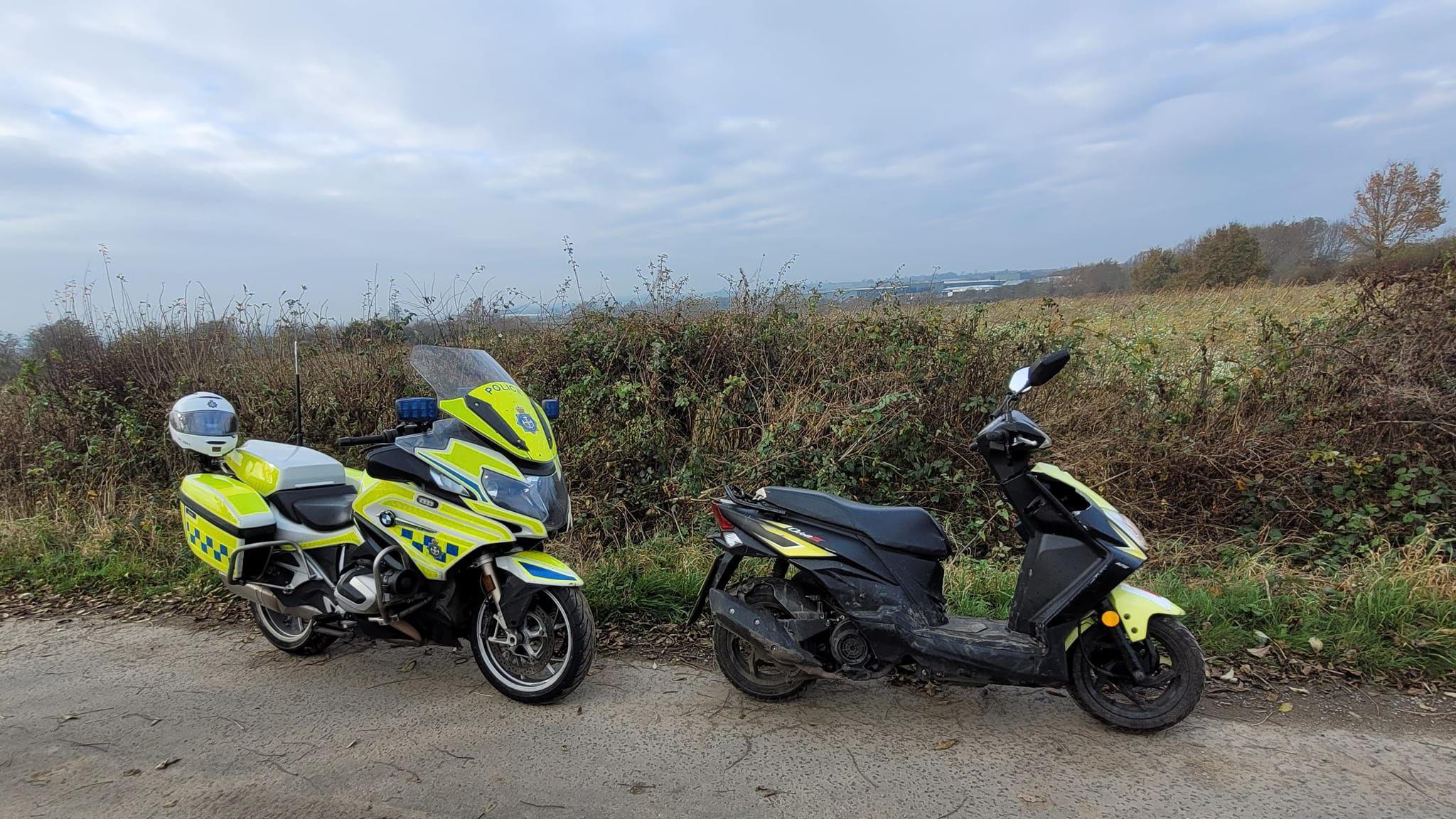 A police motorcycle and a stolen moped scooter. Hedges and a field are in the background.