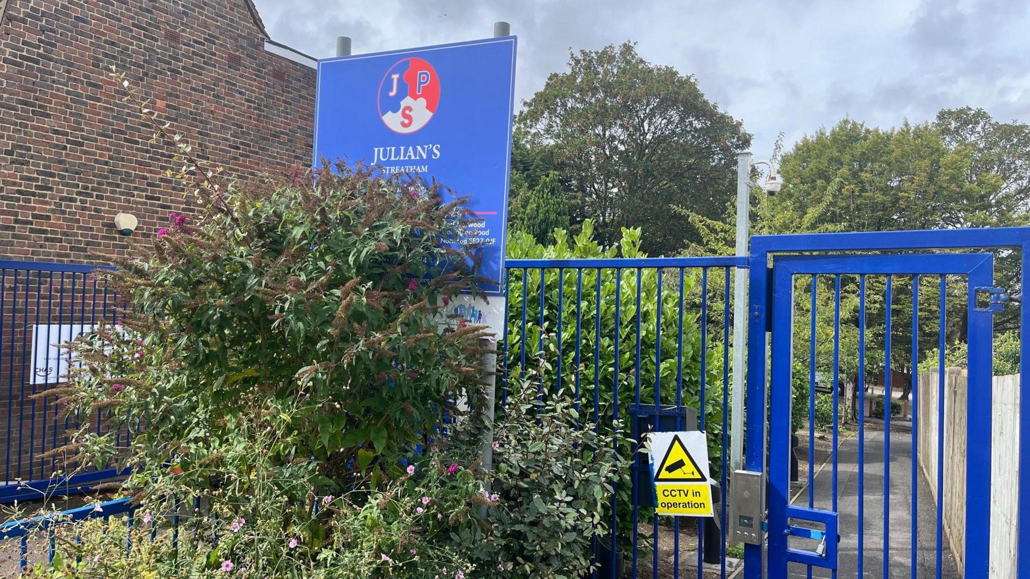 The entrance to Julian's Primary School, marked by a blue gate and a sign with the school's name and logo. In front of the gate are plants and flowers, with a brick building and trees visible in the background.