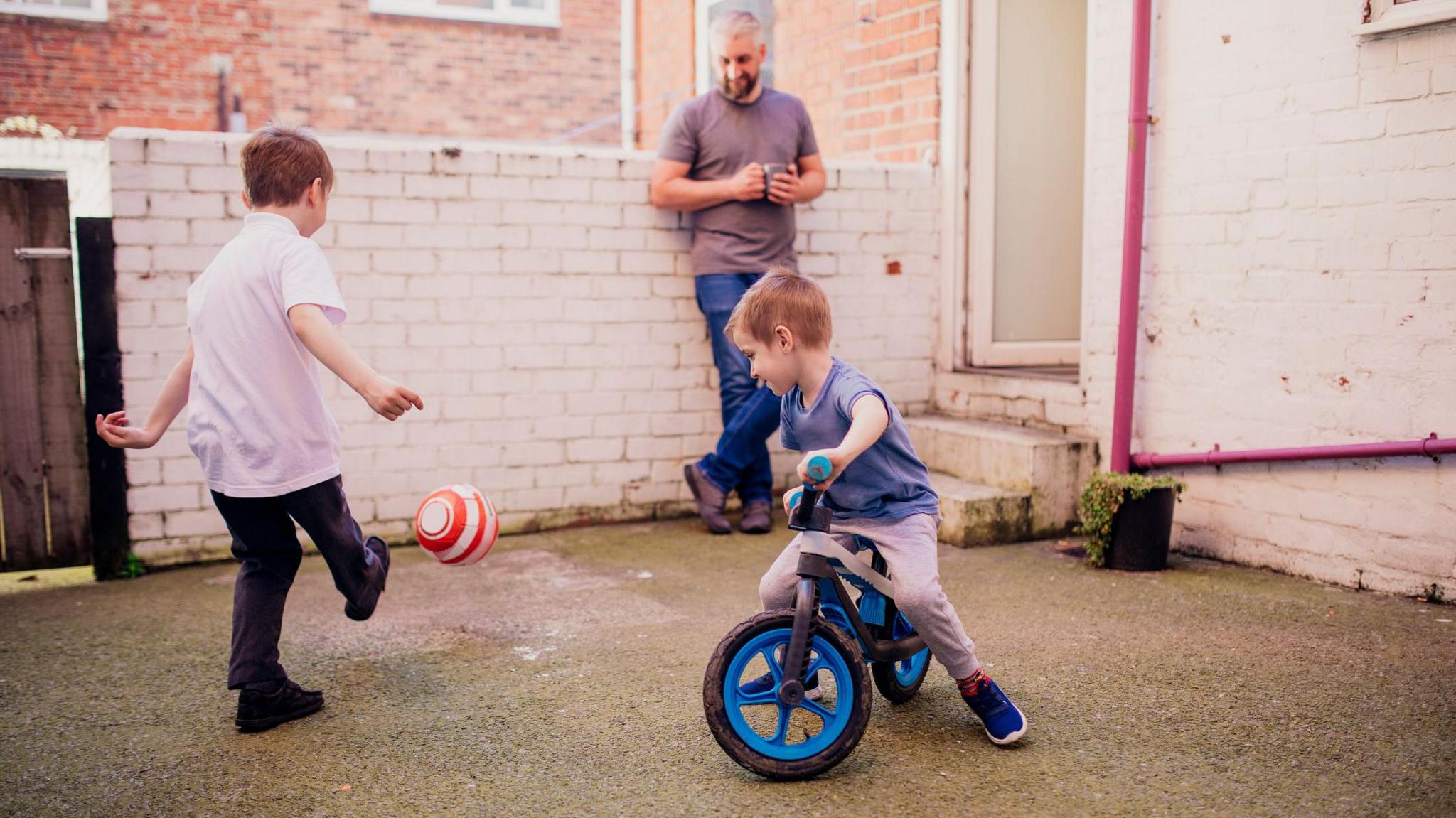 kids playing in the back yard of their house with a parent watching holding a mug of tea