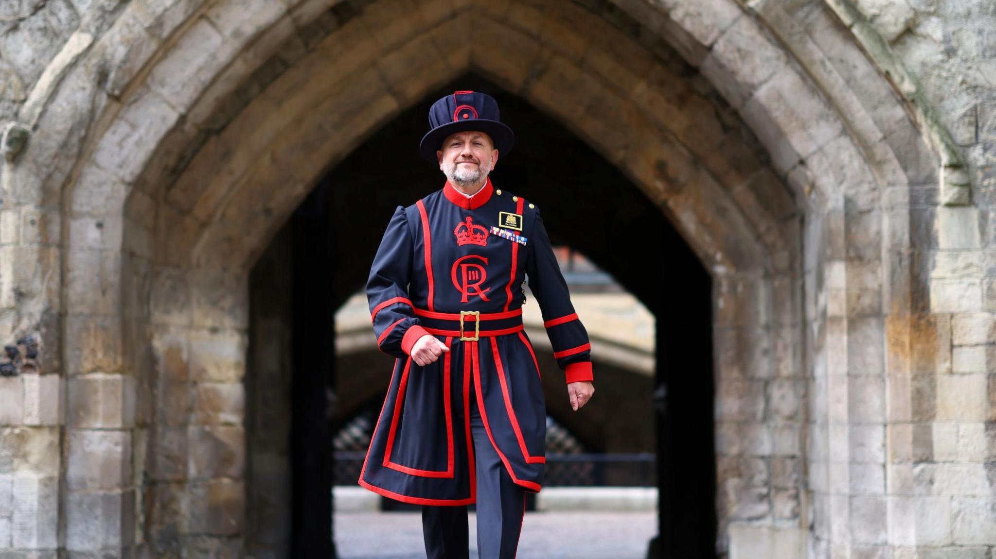 Andy Harris pictured in the grounds of the Tower of London in the daily blue and red YW uniform. He swings his arms and smiles at the camera. 
